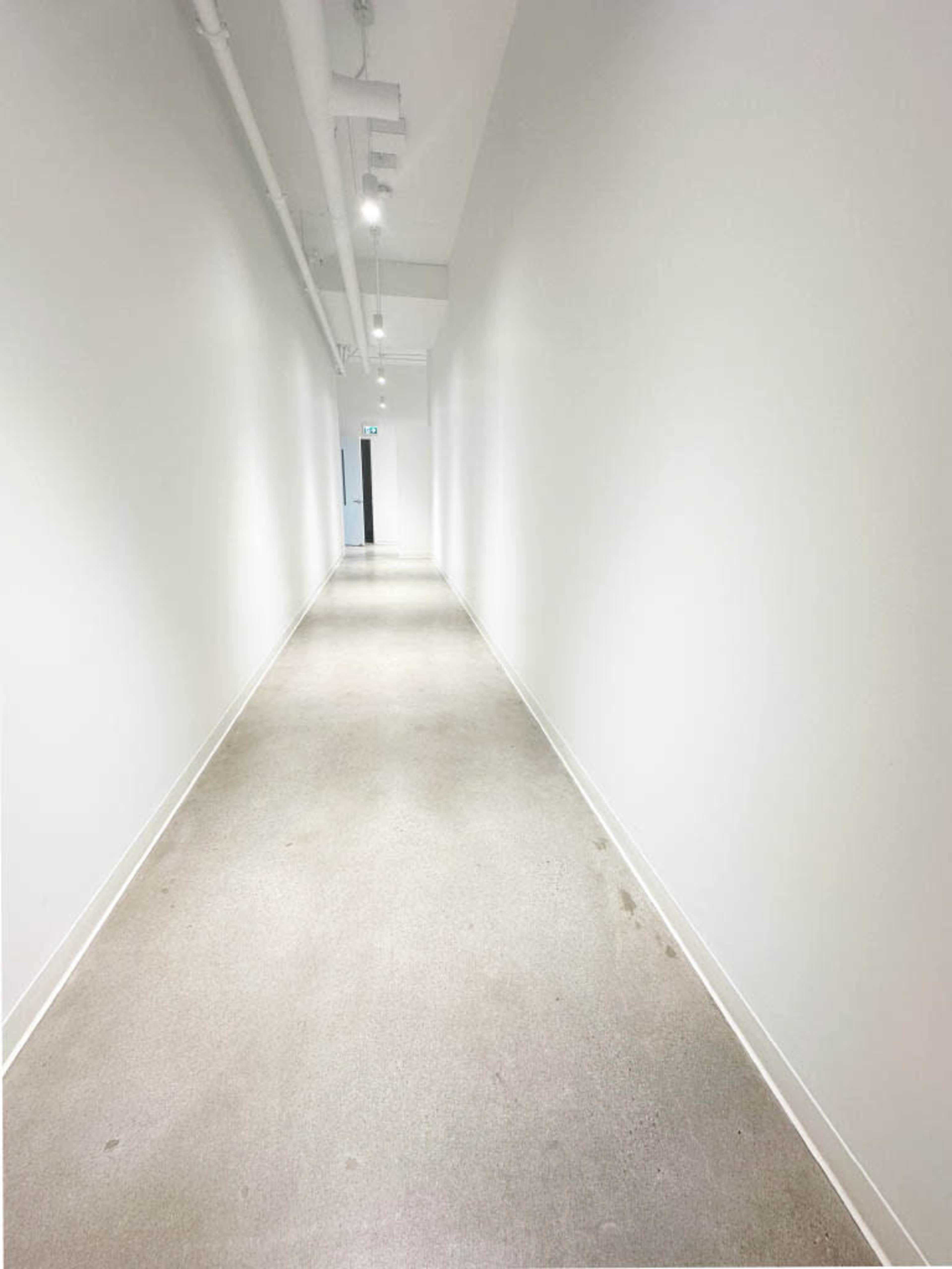 A long, narrow hallway with white walls and a polished concrete floor leads to a dark doorway at the end.