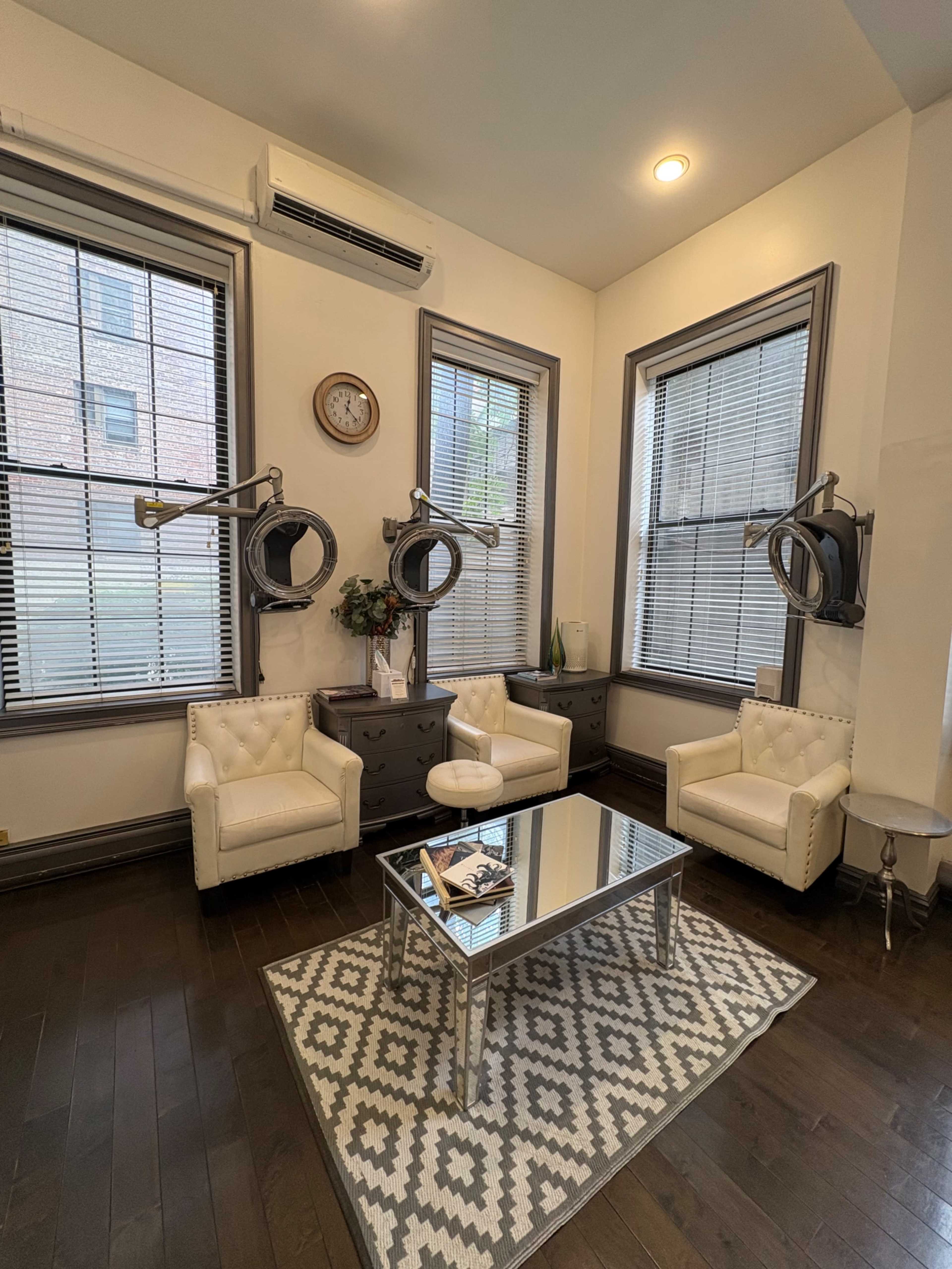 The image shows a well-furnished, contemporary room with two white armchairs, a glass coffee table on a patterned rug, and large windows featuring blinds.
