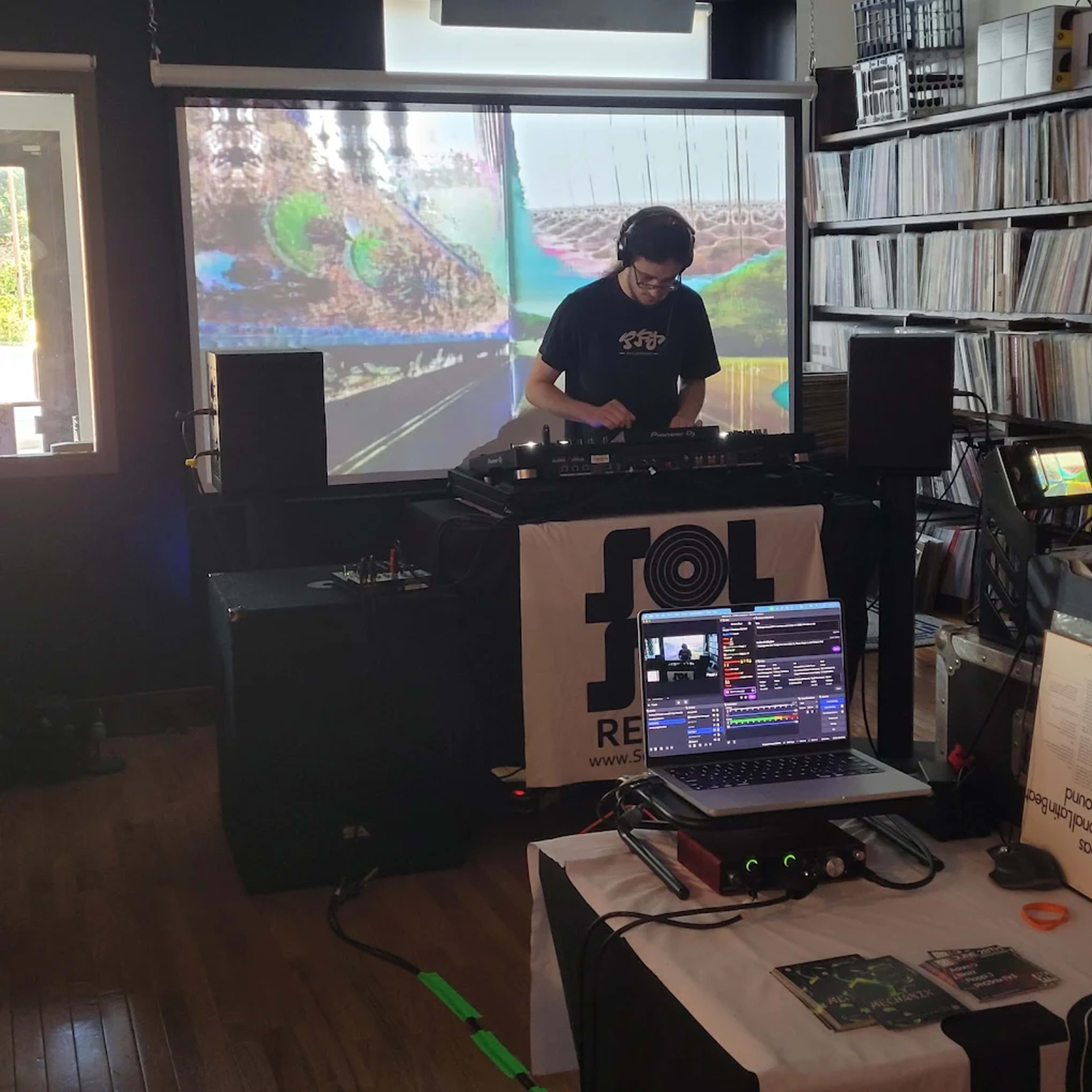 A DJ performs in front of a projection screen while surrounded by music equipment and vinyl records.