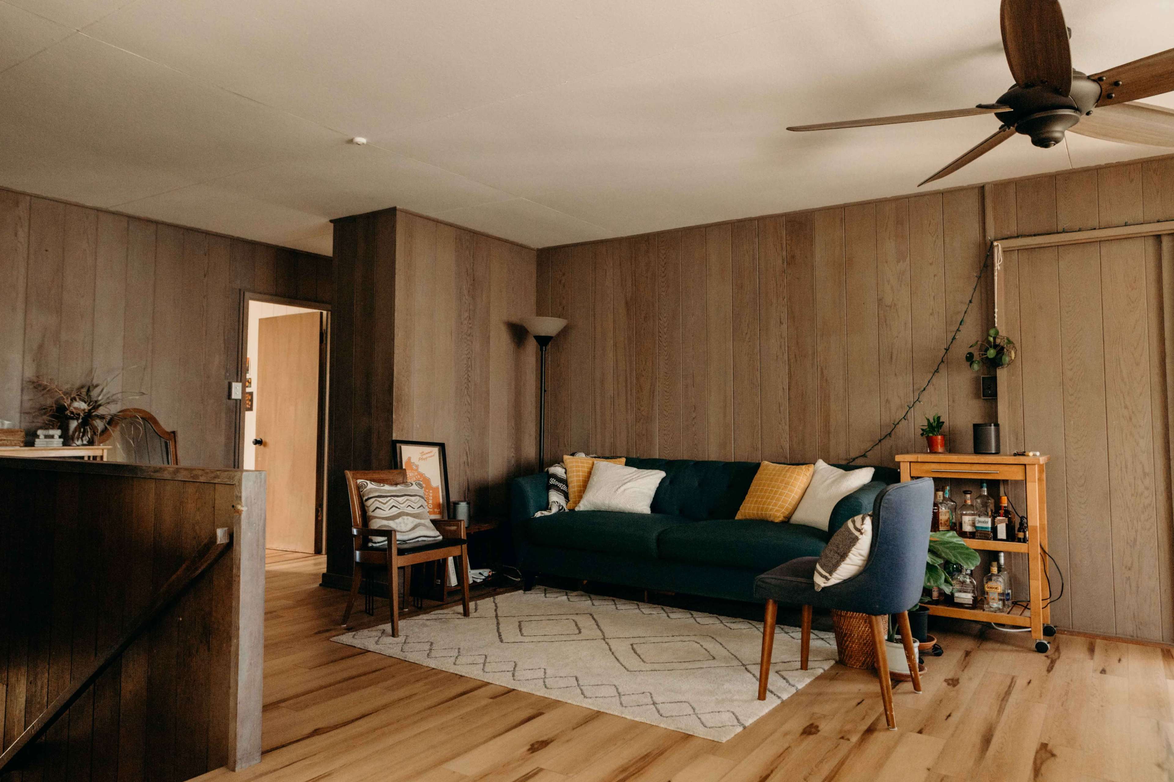 The room features a blue sofa with yellow and white pillows, a wooden floor, a fan on the ceiling, and a small bar cart in the corner.