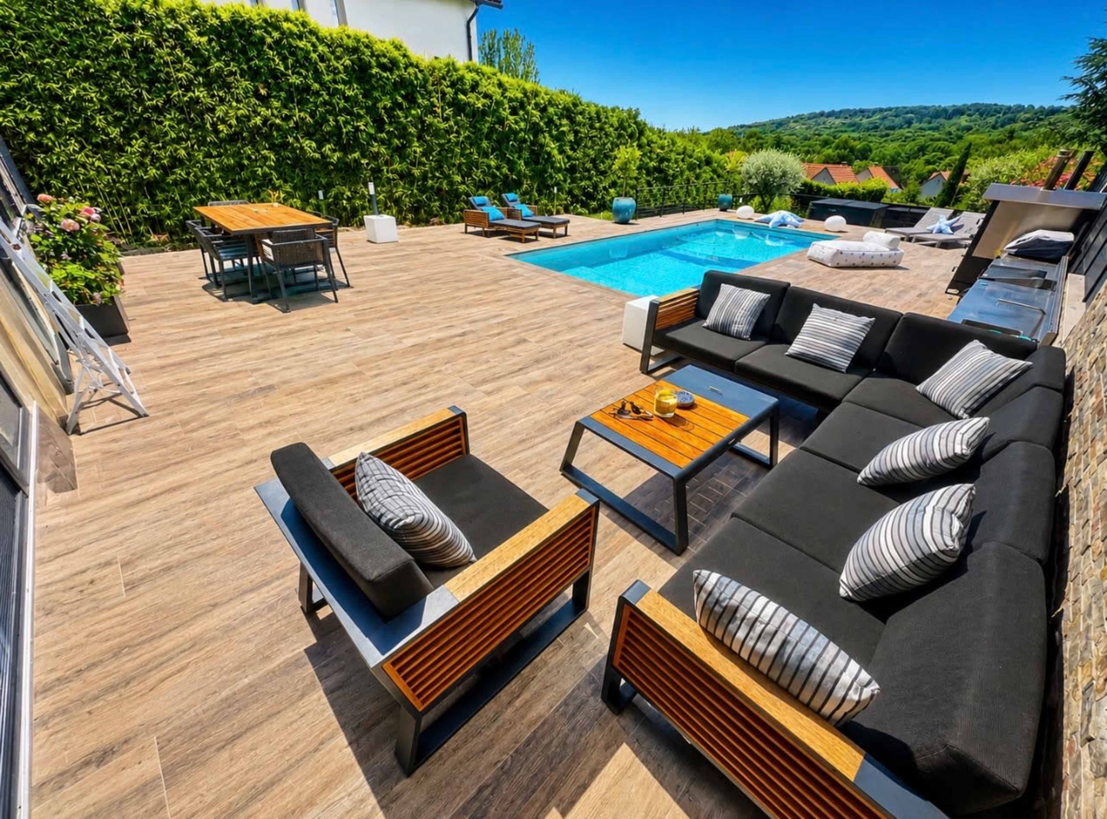 A modern outdoor space features a swimming pool surrounded by wooden decking, with stylish lounge furniture and a dining area amidst lush greenery.