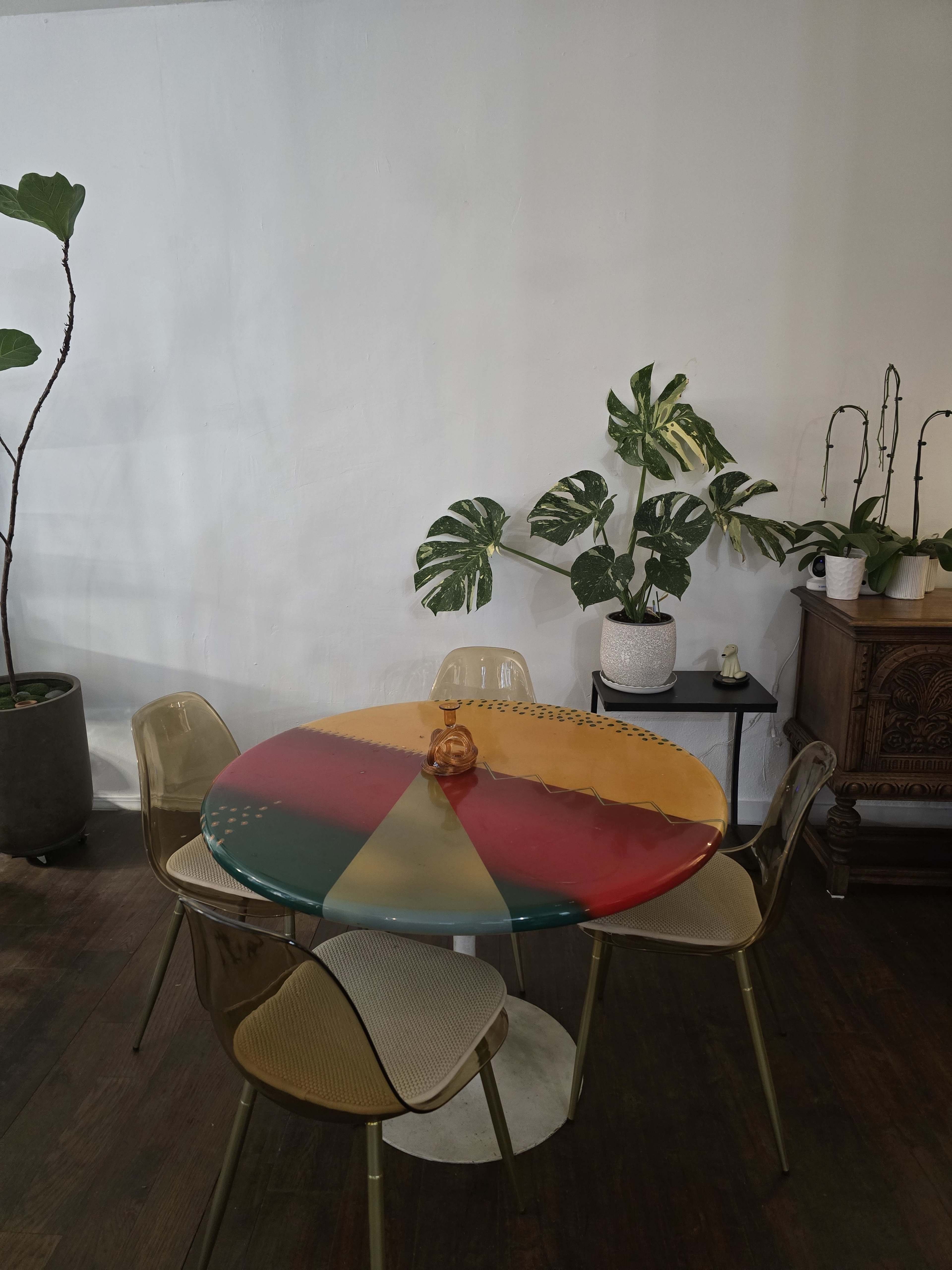 A round, multicolored table is set in a room with potted plants and wooden flooring.