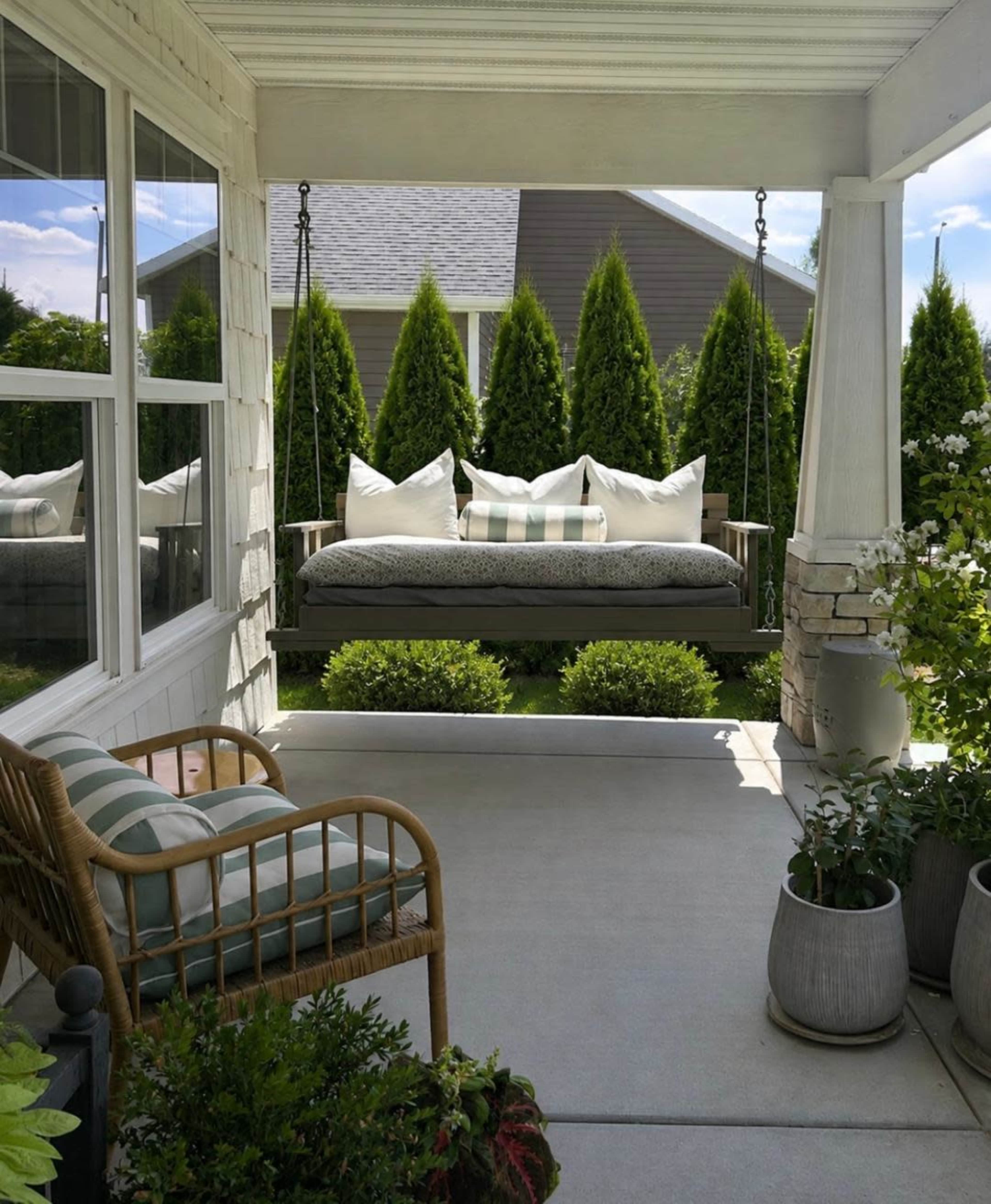 A covered porch features a swing seat with pillows and a nearby wicker chair, surrounded by well-maintained greenery and potted plants.