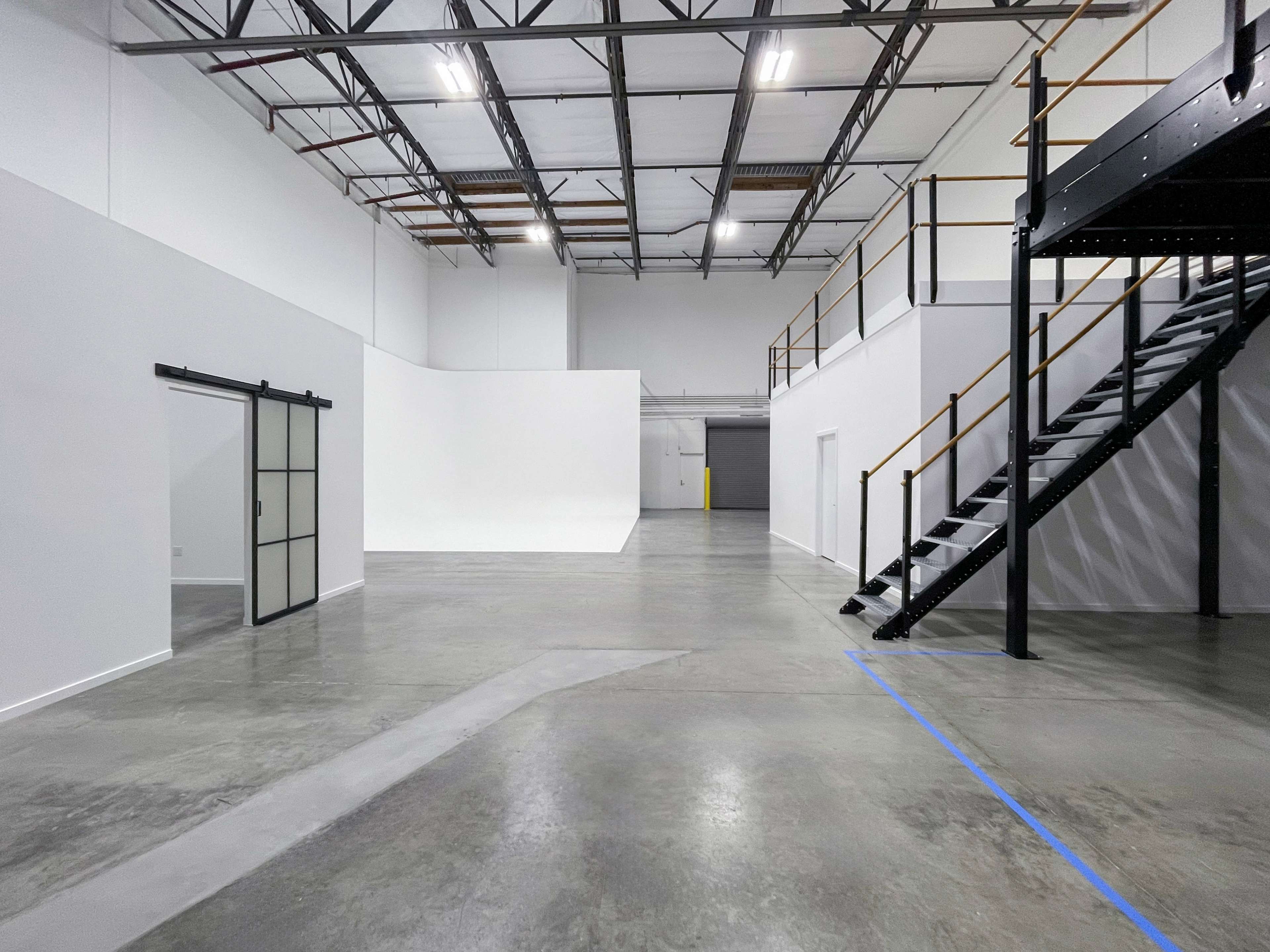 The image shows an empty industrial space with high ceilings, concrete flooring, and a staircase leading to a second level.