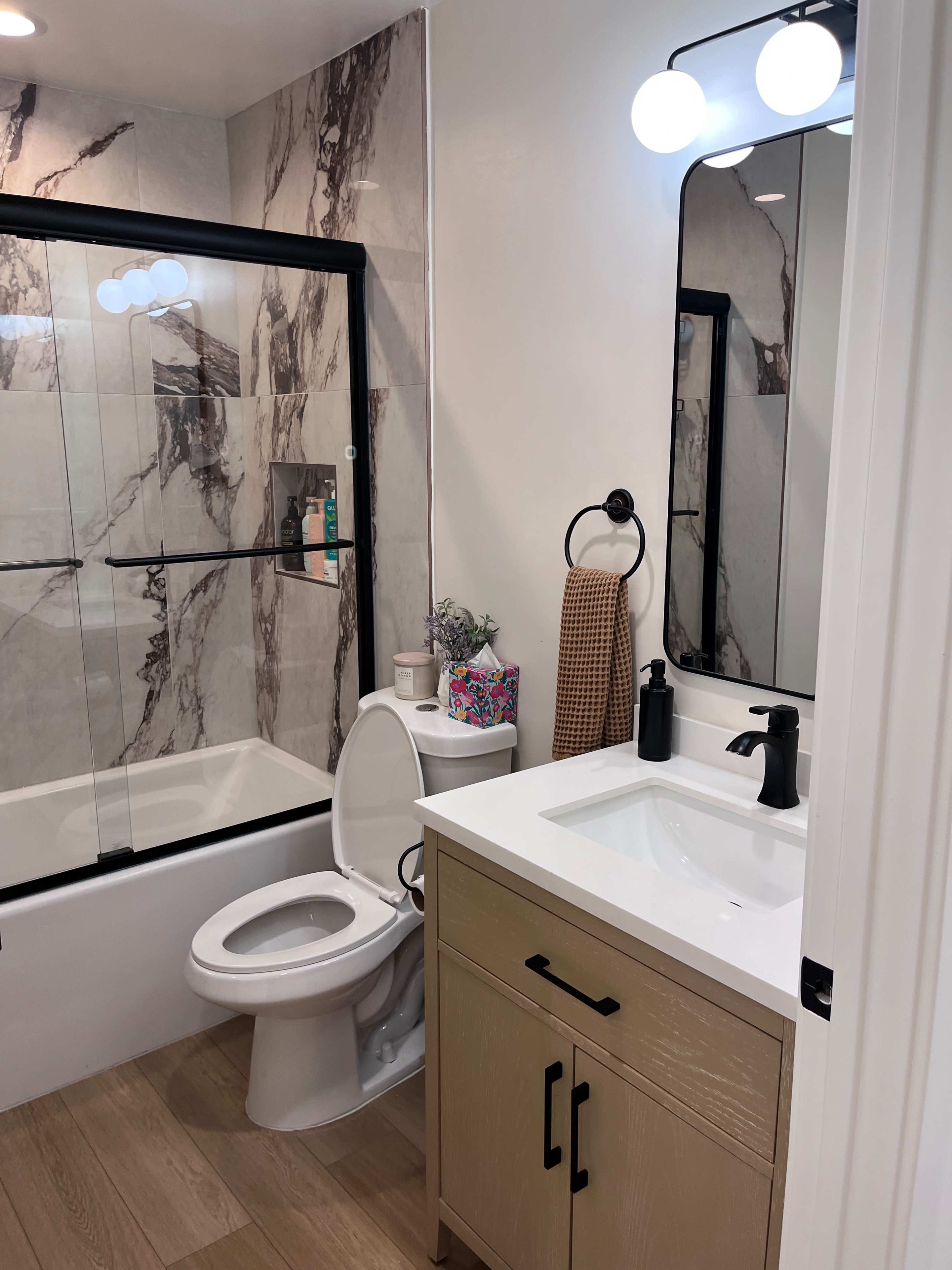 The image shows a modern bathroom featuring a glass shower enclosure, a white bathtub, a toilet, a wooden vanity with a sink, and a large mirror with stylish lighting.