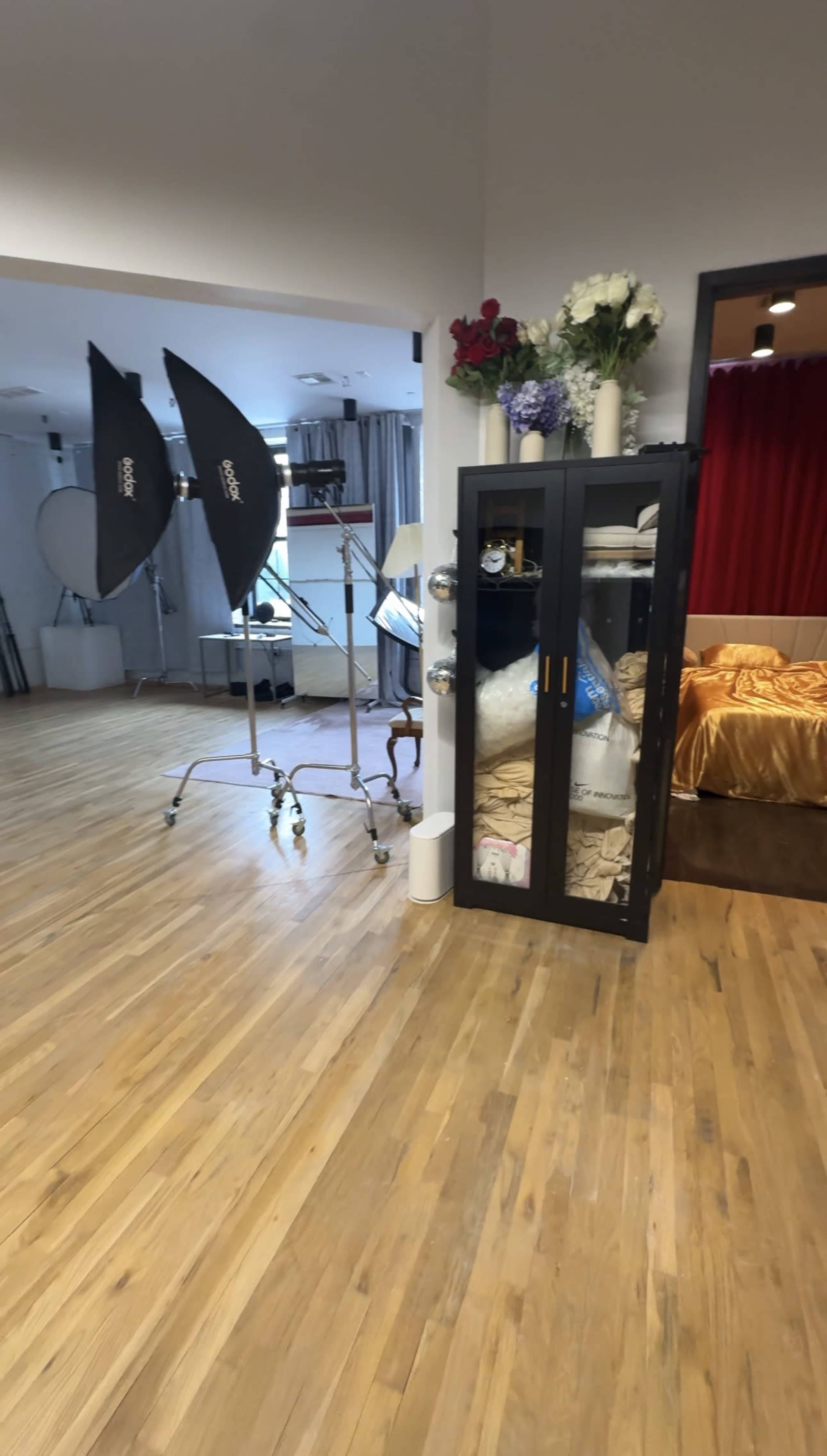 The image shows a studio space with wooden flooring, two large light umbrellas on stands, and a glass-fronted cabinet next to a decorated bed.