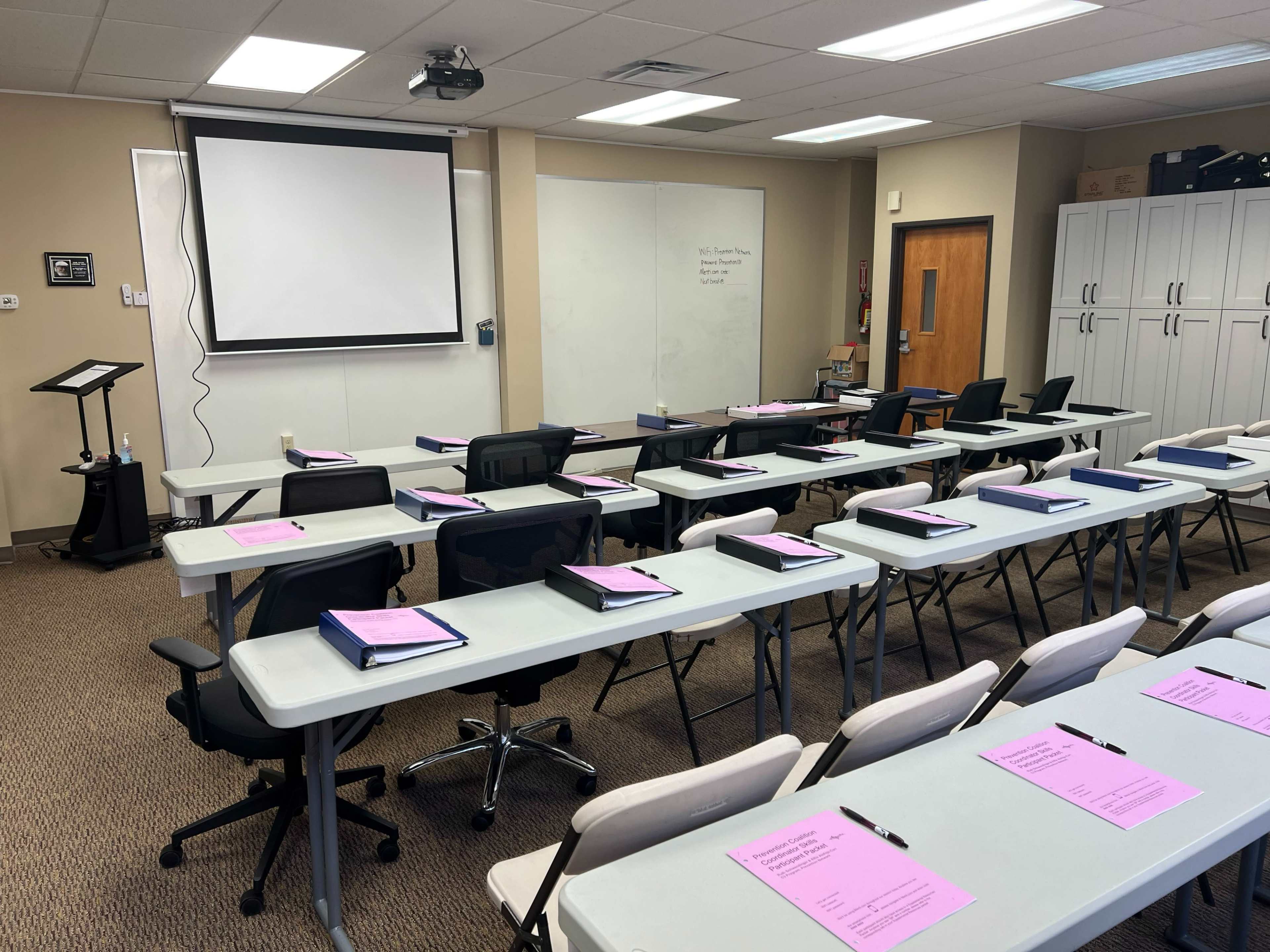 A classroom setting with rows of desks arranged in front of a projector screen, each desk featuring a pink folder and a pen.