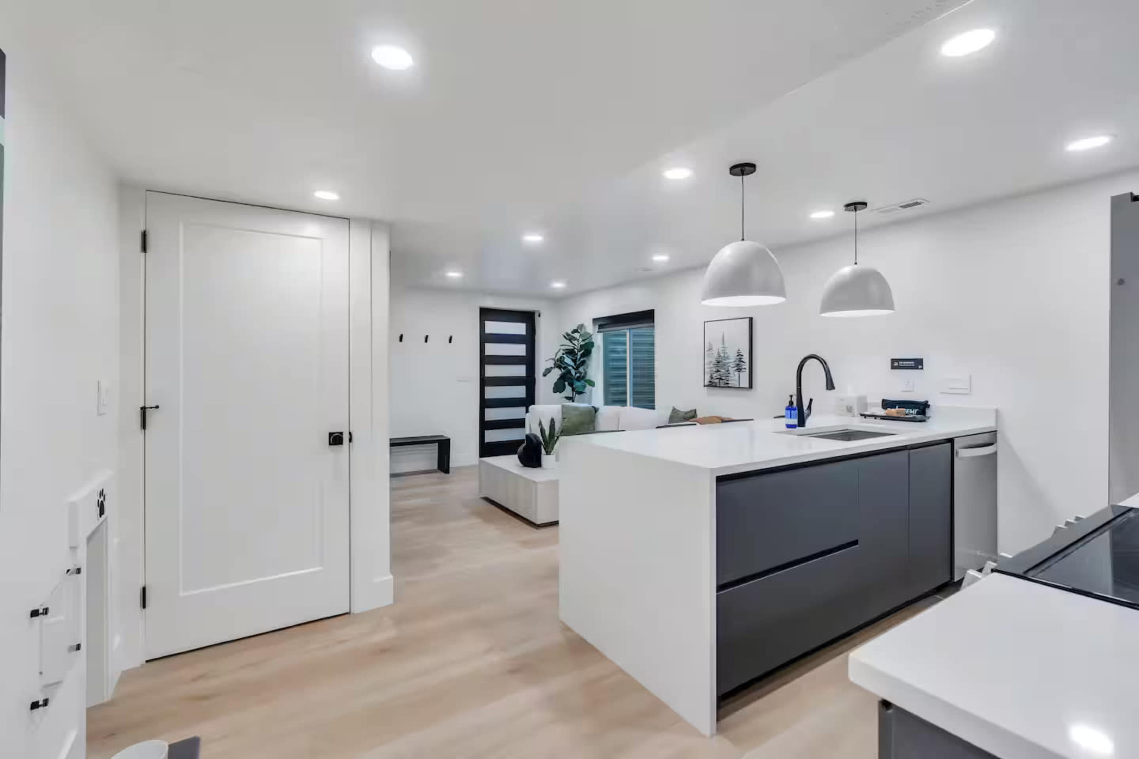 The image shows a modern kitchen with a white island, dark cabinetry, and pendant lights, connected to a light-filled living area.