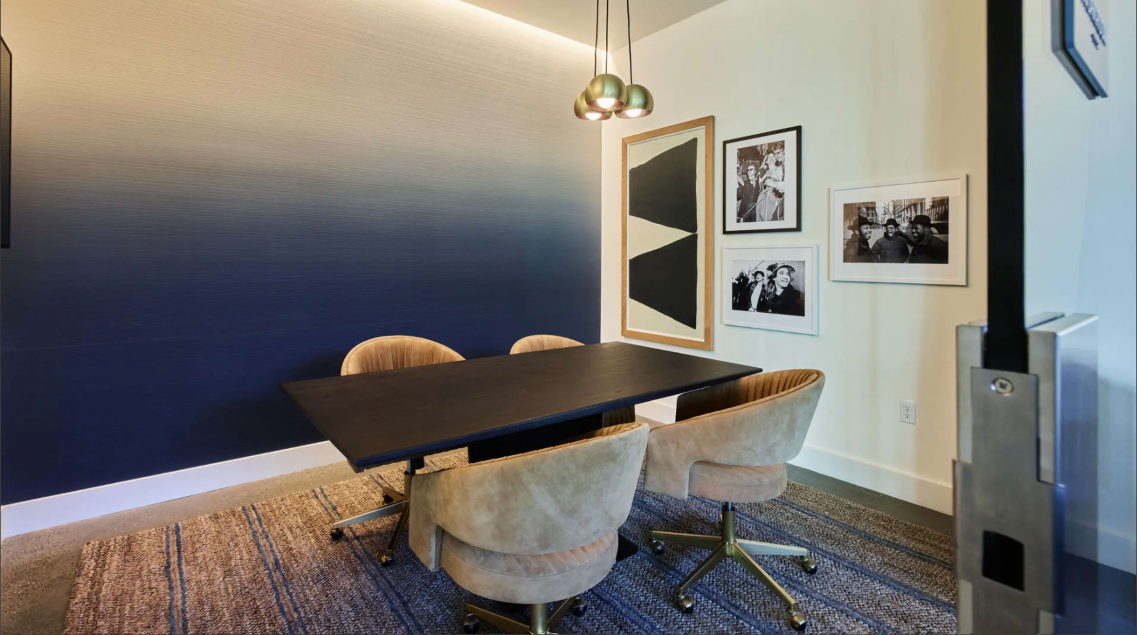 The image shows a small meeting room featuring a dark table surrounded by four upholstered chairs, with a gradient blue wall and framed artwork on one side.
