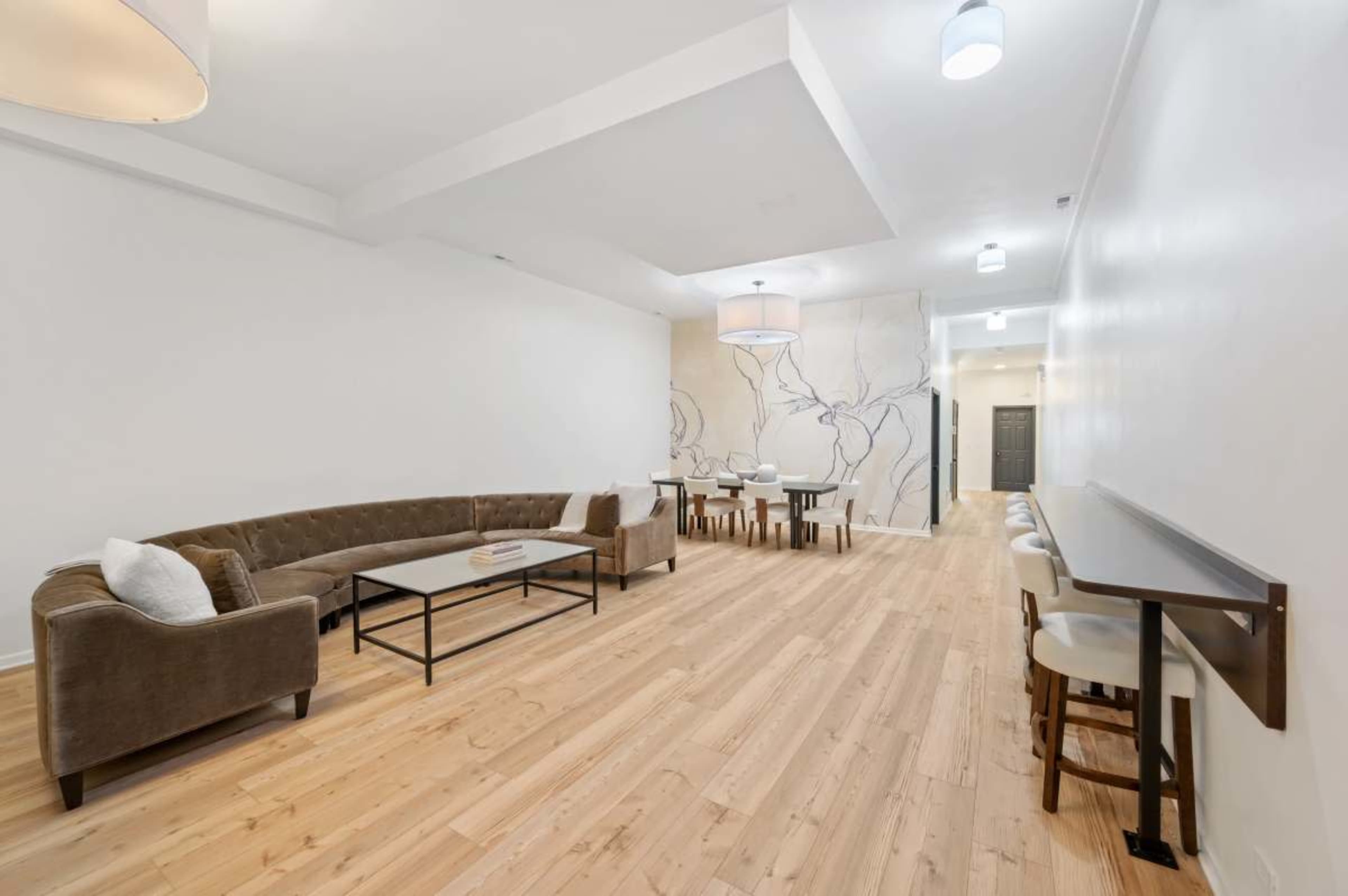 The image shows a spacious, modern interior with light wood flooring, a large sectional sofa, a coffee table, a dining area with several chairs, and decorative wall art.