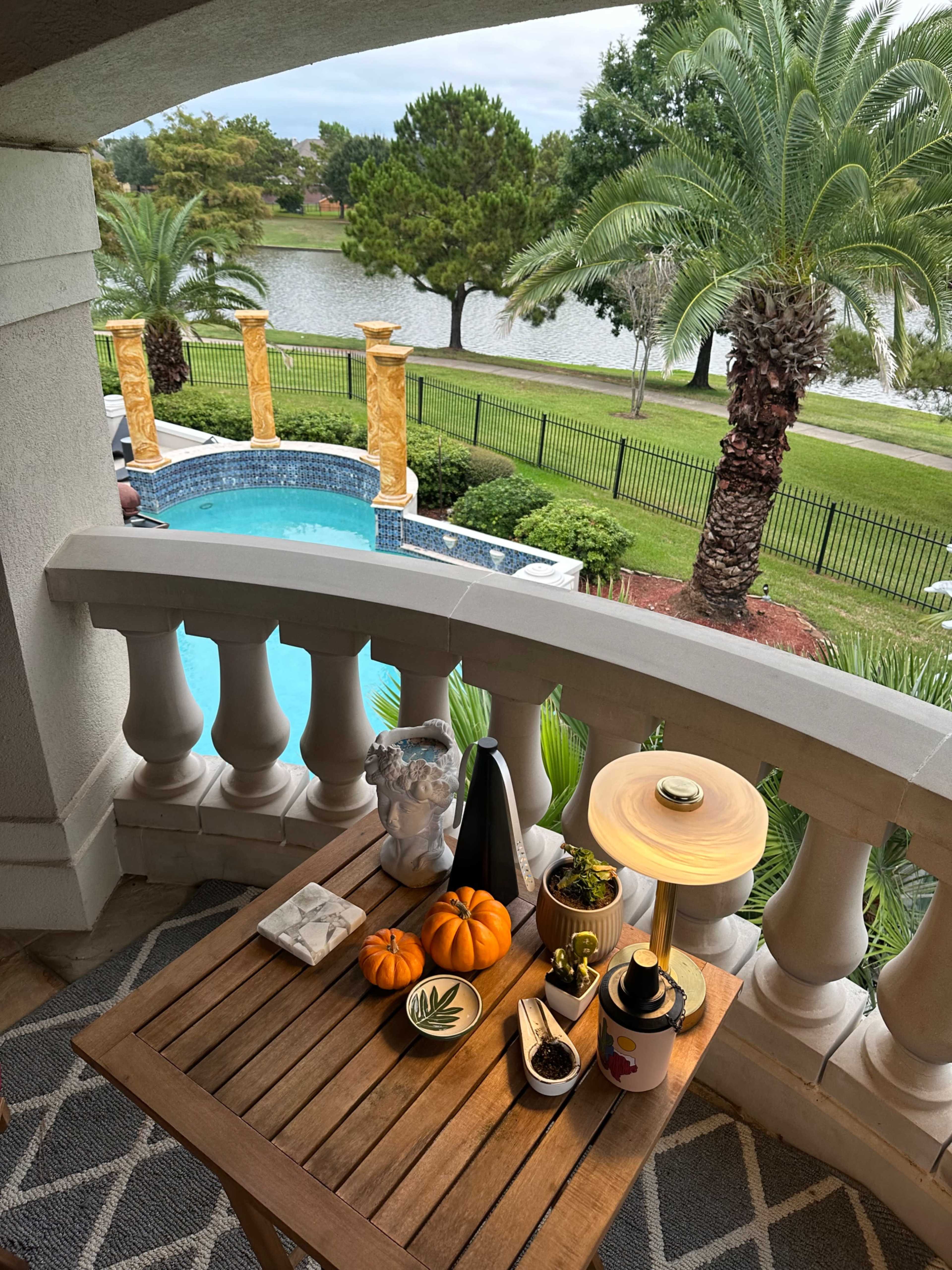 A wooden table on a balcony displays decorative items, including pumpkins and a lamp, overlooking a pool and a lake surrounded by palm trees.