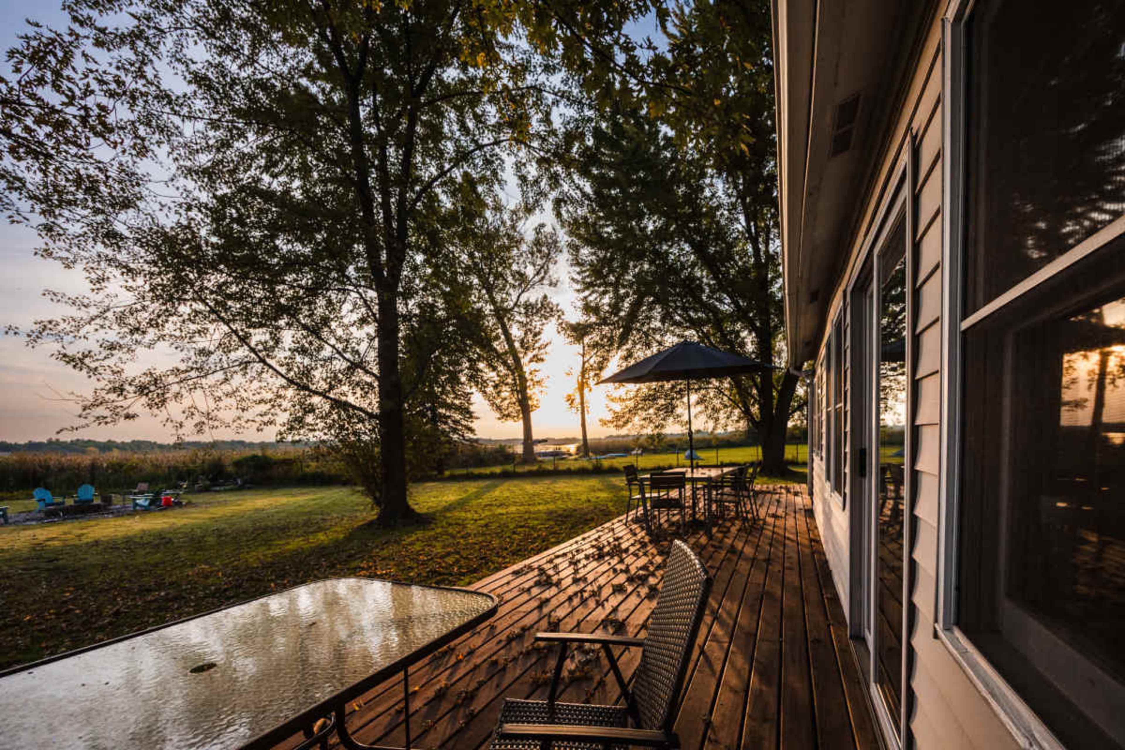The image shows a wooden deck next to a house, with chairs and a table, overlooking a grassy area and a sunset in the background.