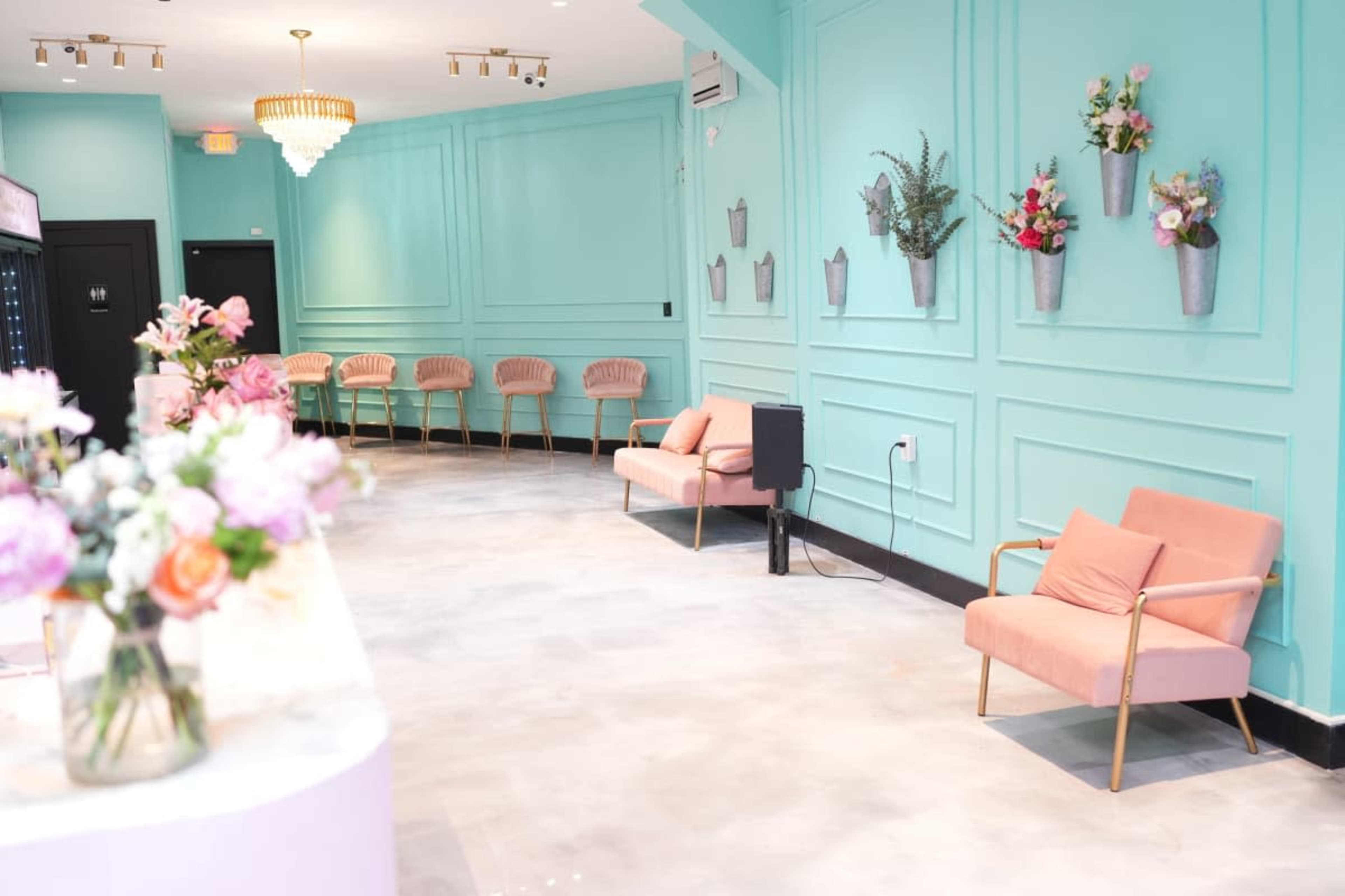 A brightly colored room features a mint green wall with floral wall hangings and two pink upholstered chairs opposite a reception area adorned with a floral arrangement.