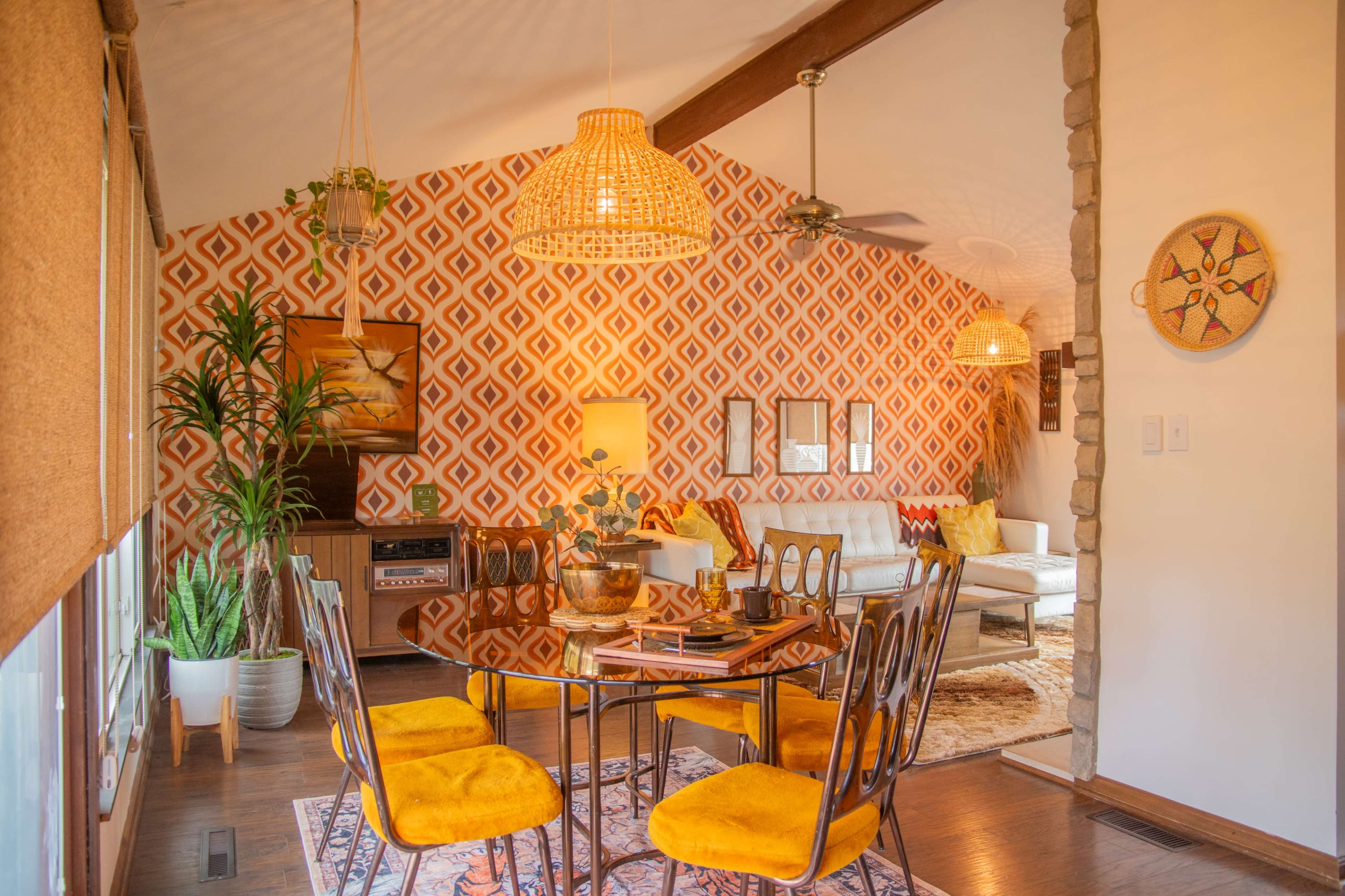 The image shows a retro-style dining area with a round table surrounded by bright yellow chairs, set against a patterned orange wall and featuring decorative plants and warm lighting.