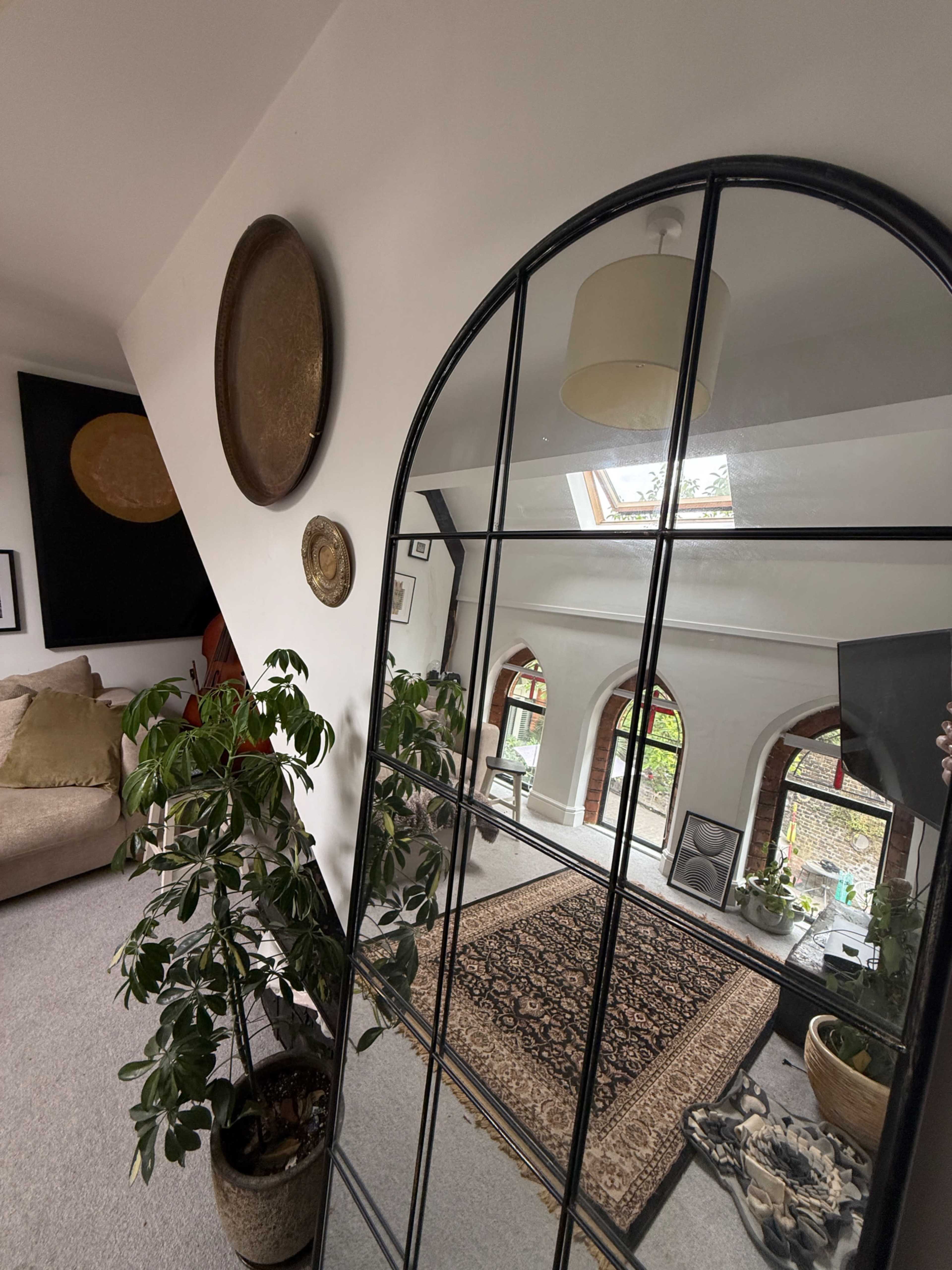 A large arched mirror reflects a living room with a sofa, plants, and a decorative rug.