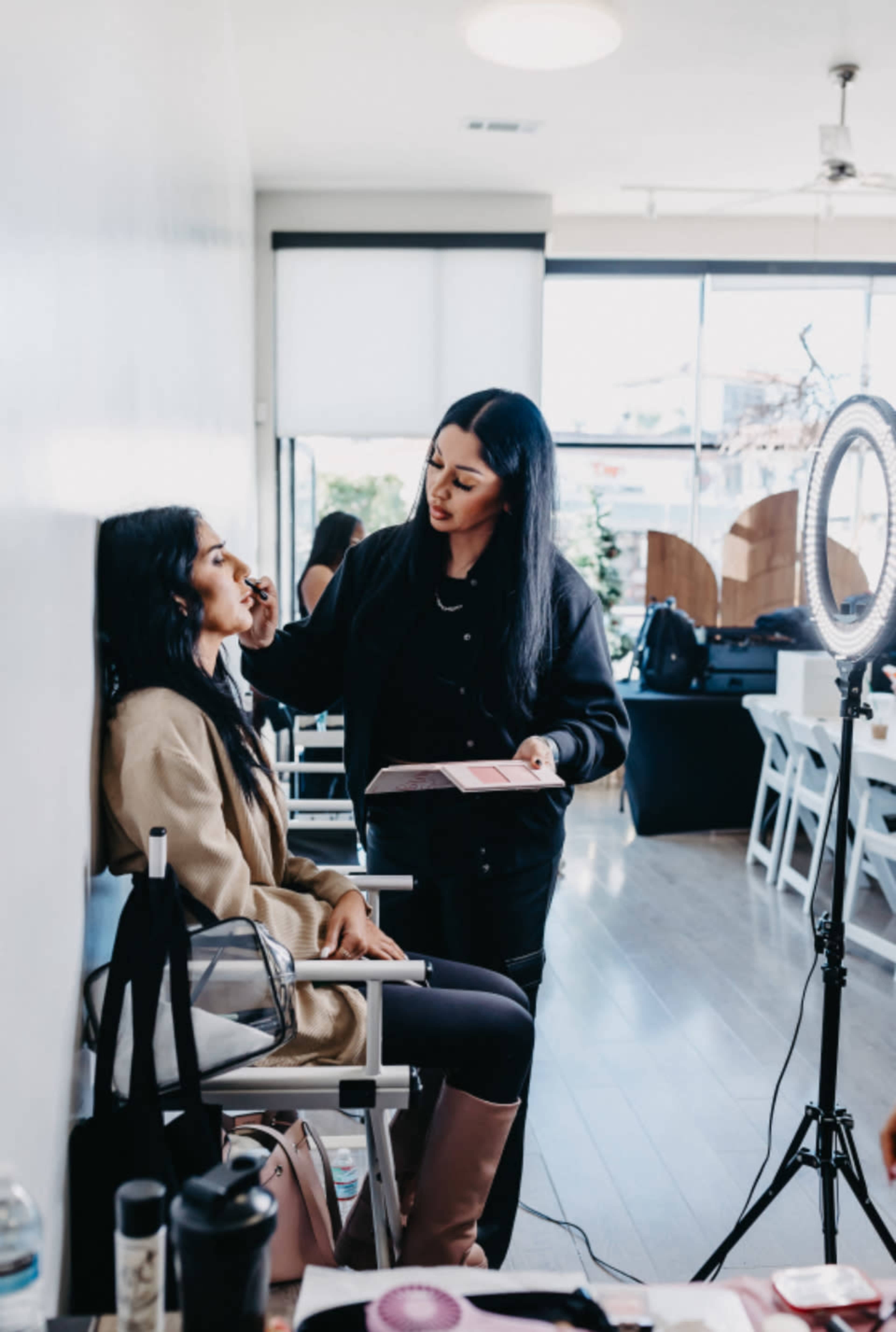 A makeup artist applies cosmetics to a seated client in a well-lit studio with white walls.
