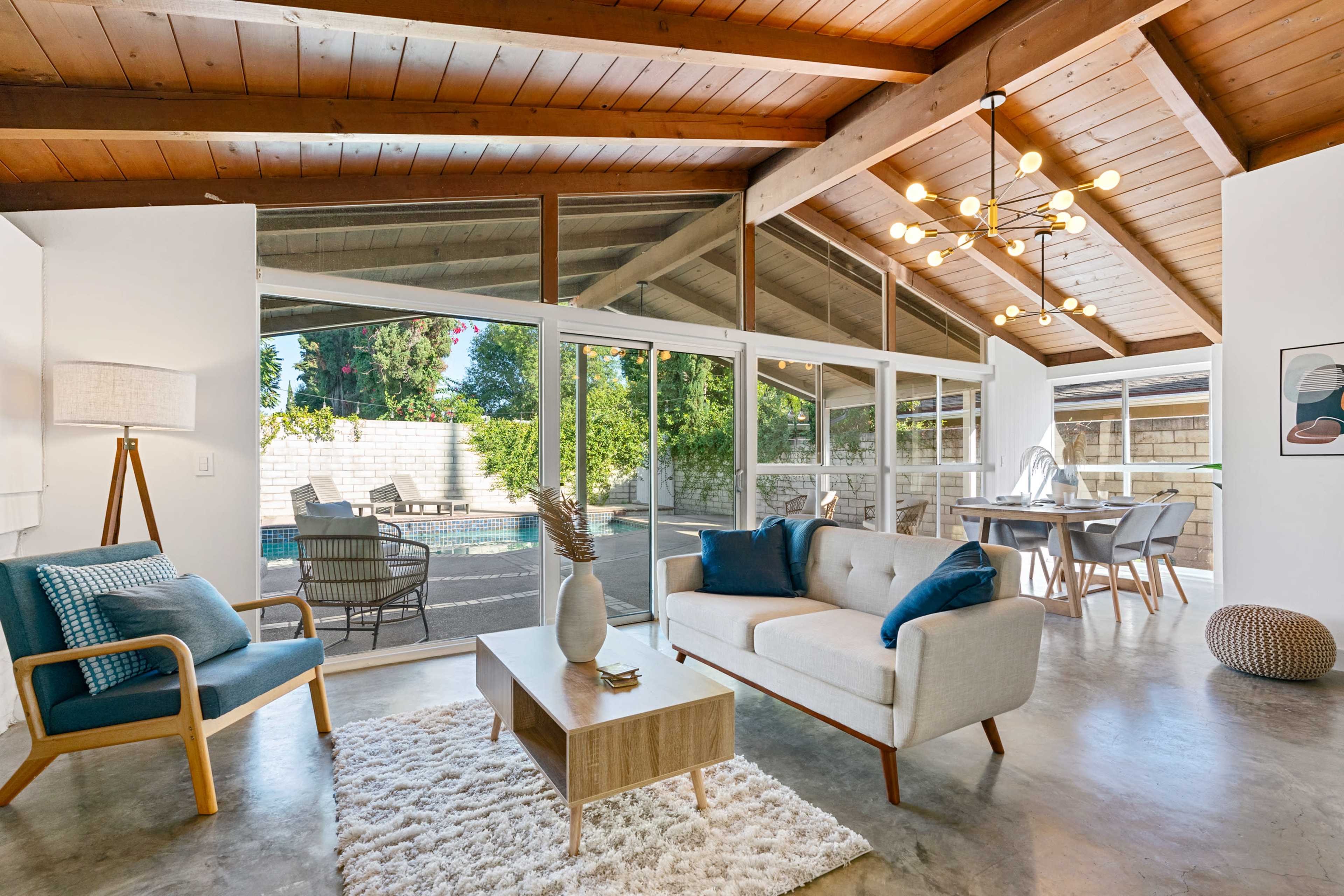A modern living room features a large glass wall overlooking a patio and pool, with wooden beams and a minimalist decor.