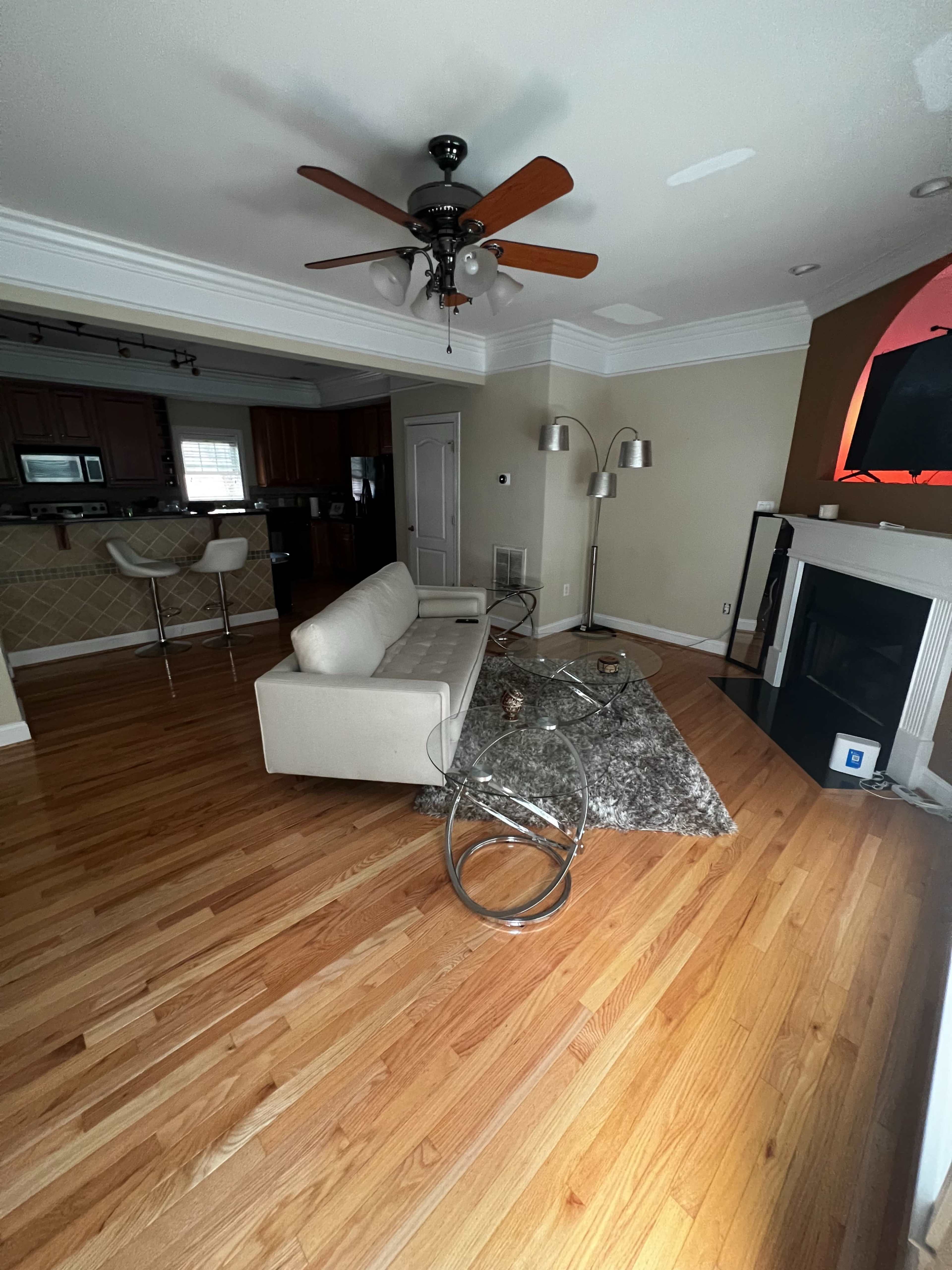 A living room with a white sofa, a glass coffee table, and a fireplace, featuring wooden flooring and a ceiling fan.