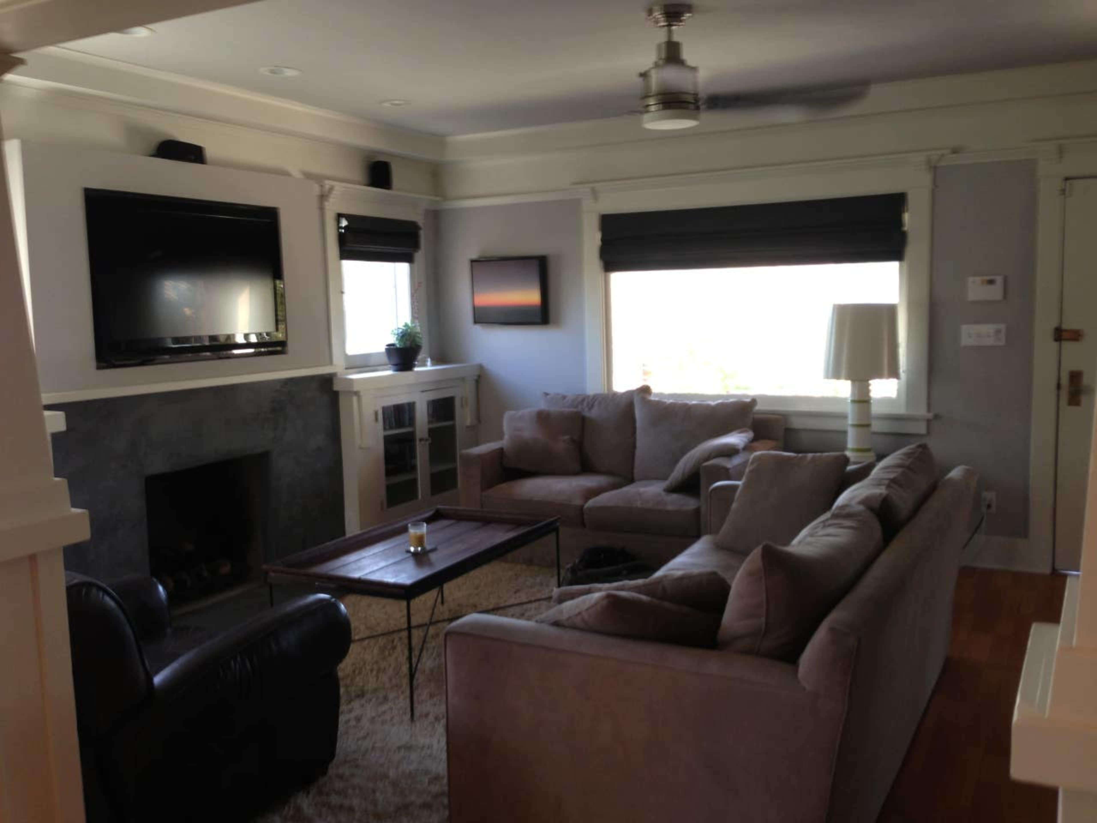 A living room with a sectional sofa, a coffee table, a television mounted on the wall, and large windows with roller shades.