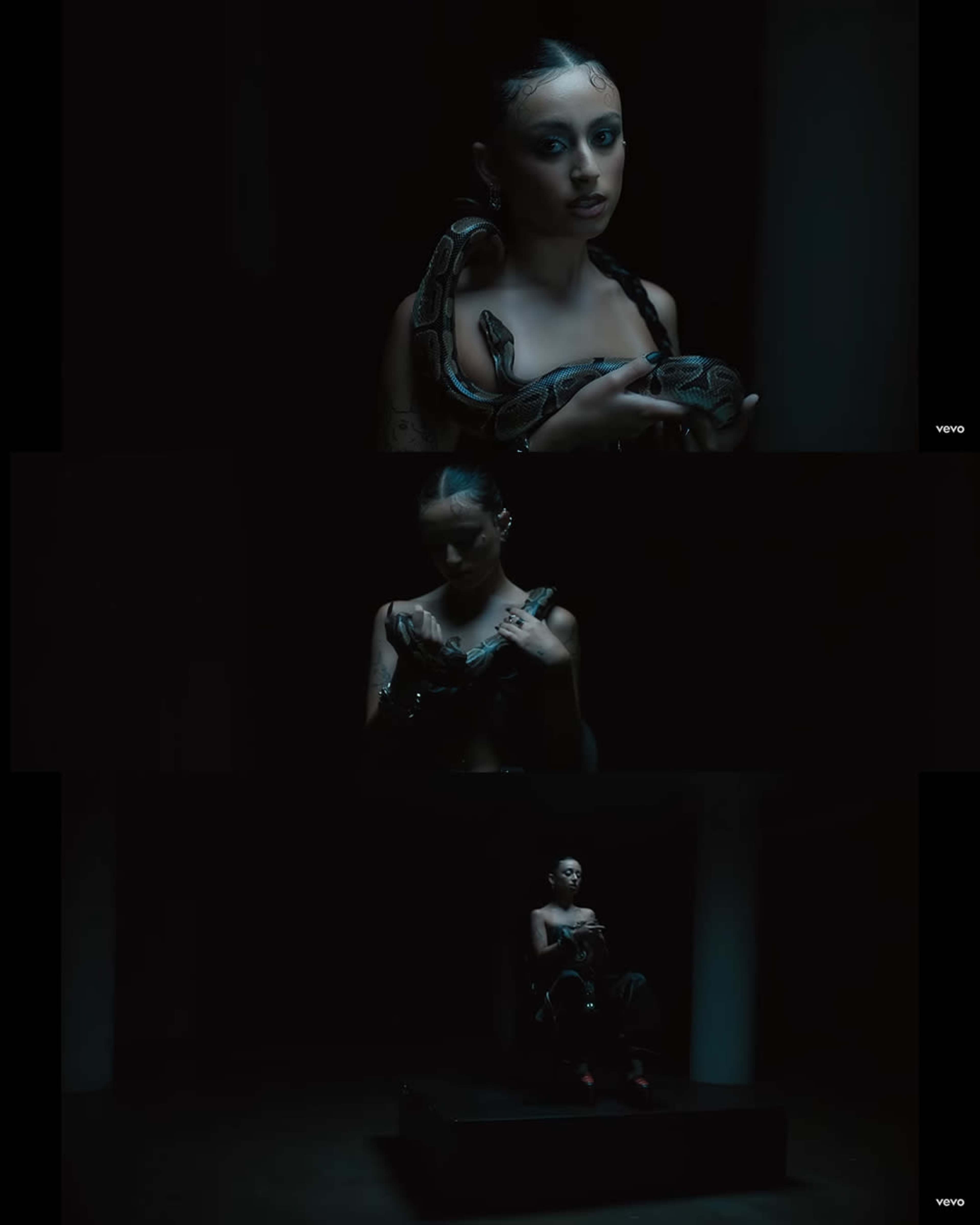 A woman with long hair holds a snake while seated in a dimly lit room.