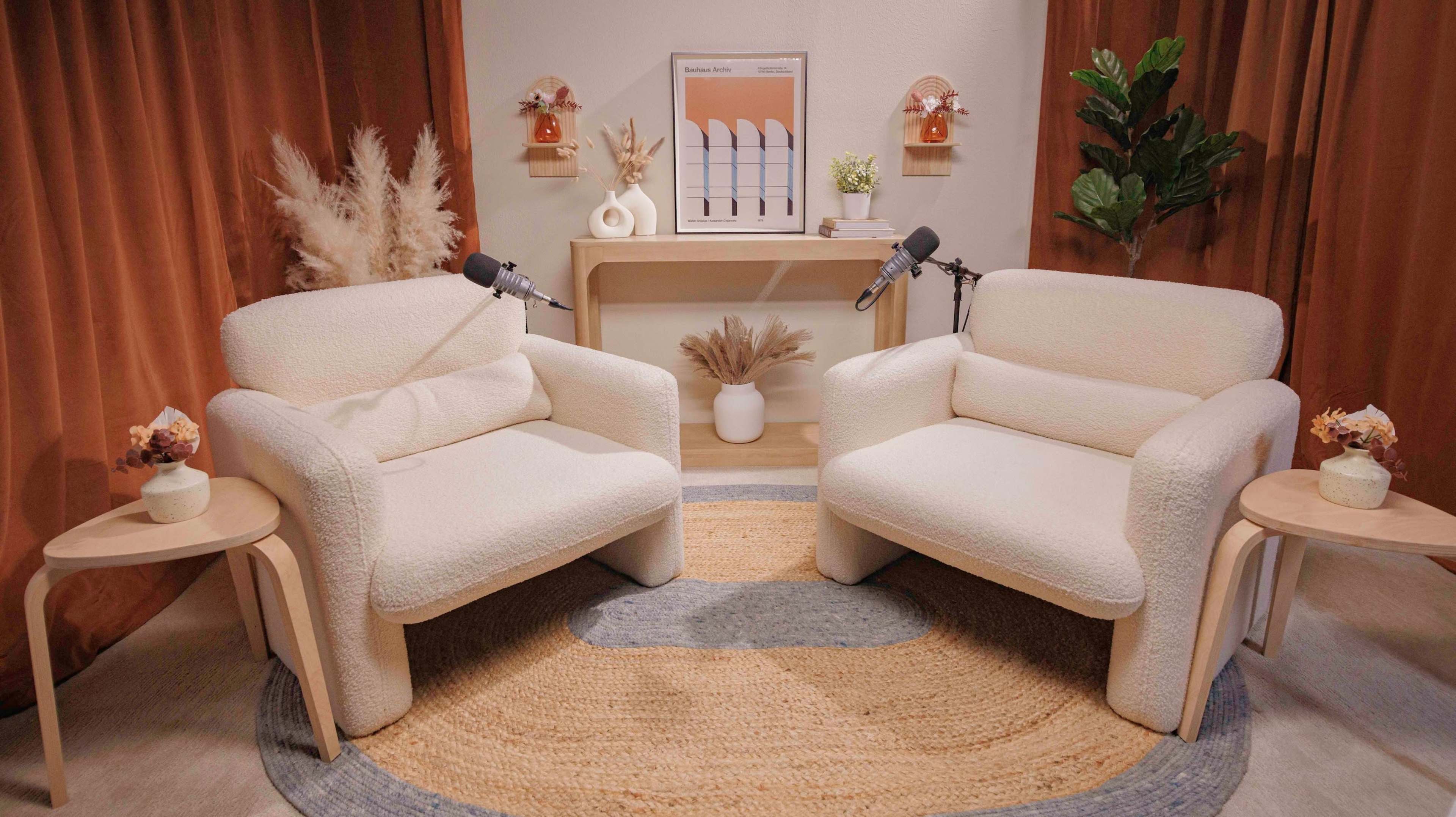 The scene displays a cozy living area featuring two white, textured armchairs facing each other, with a round rug beneath, accompanied by small side tables and decorative plants.