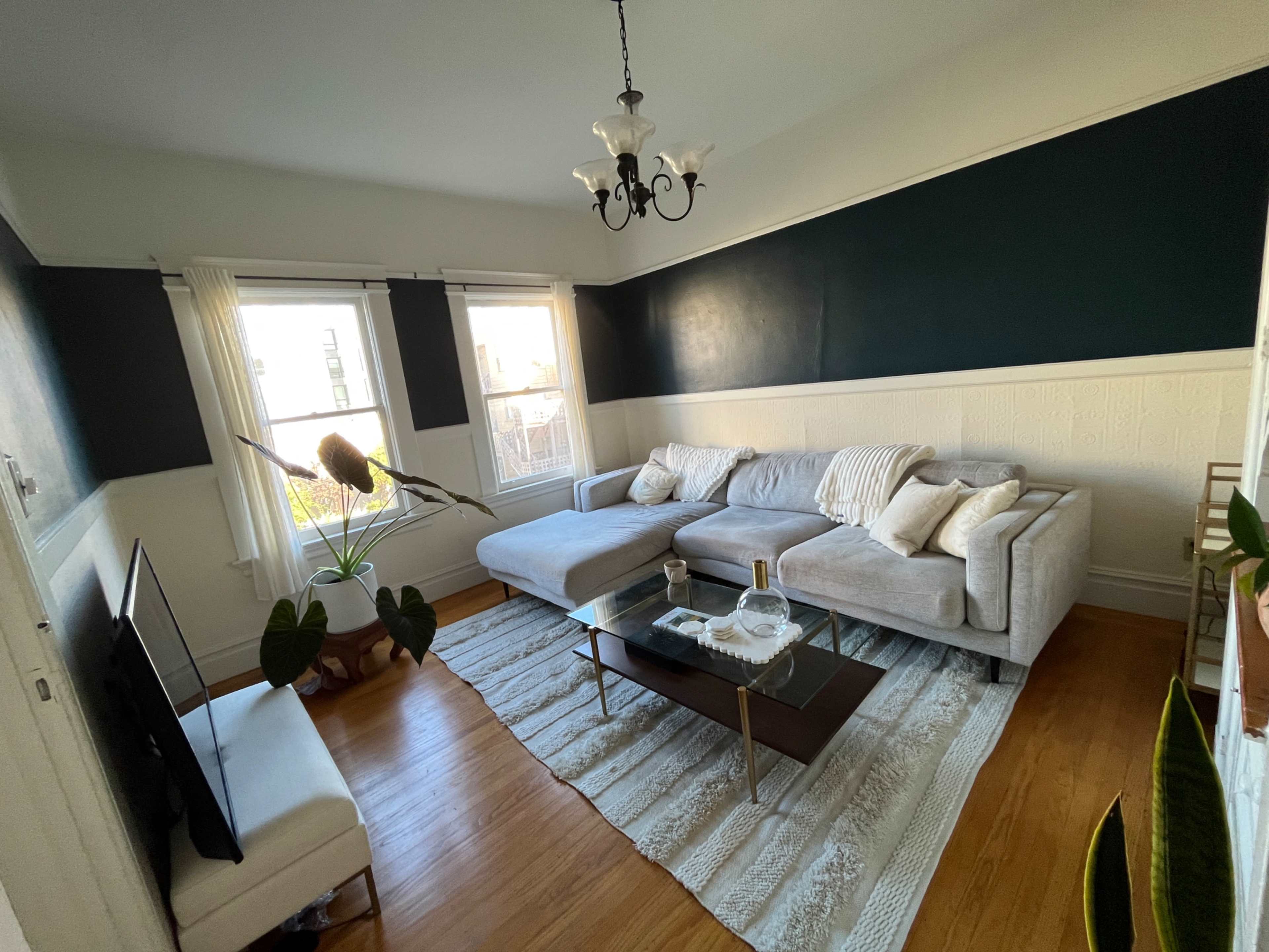 A minimalist living room features a gray sectional sofa, a coffee table, large windows, and decorative plants.