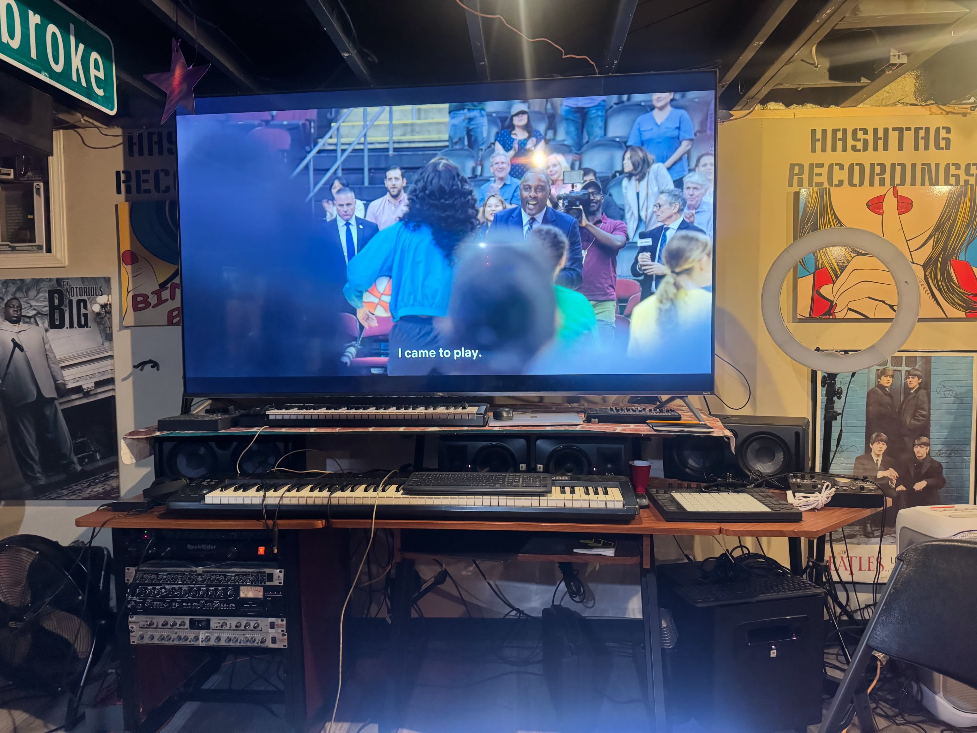 A large television displays a scene from a sports-themed show in a basement filled with music equipment and posters.