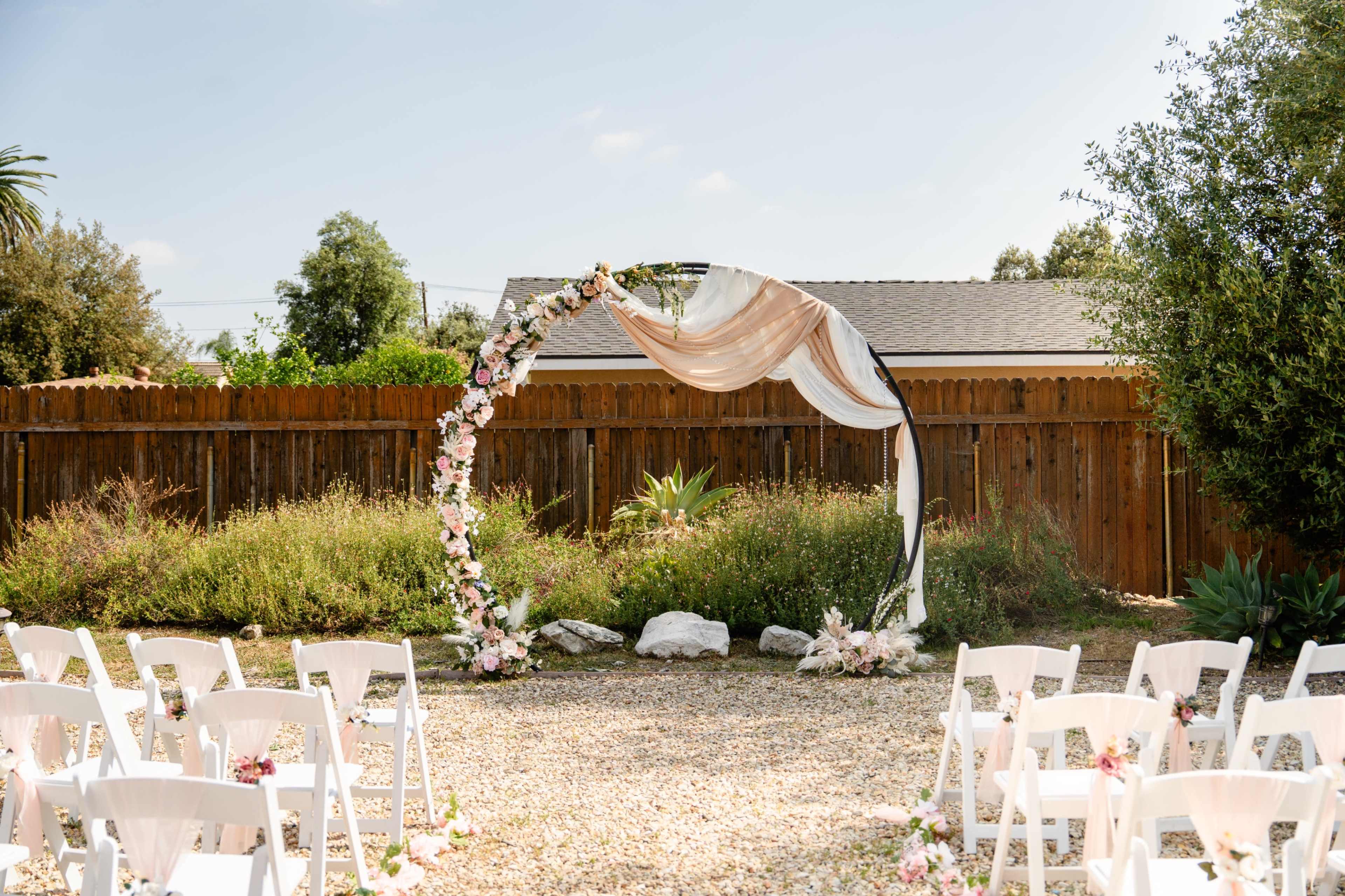 A wedding altar with floral decorations and draped fabric is set up in a garden surrounded by white folding chairs.
