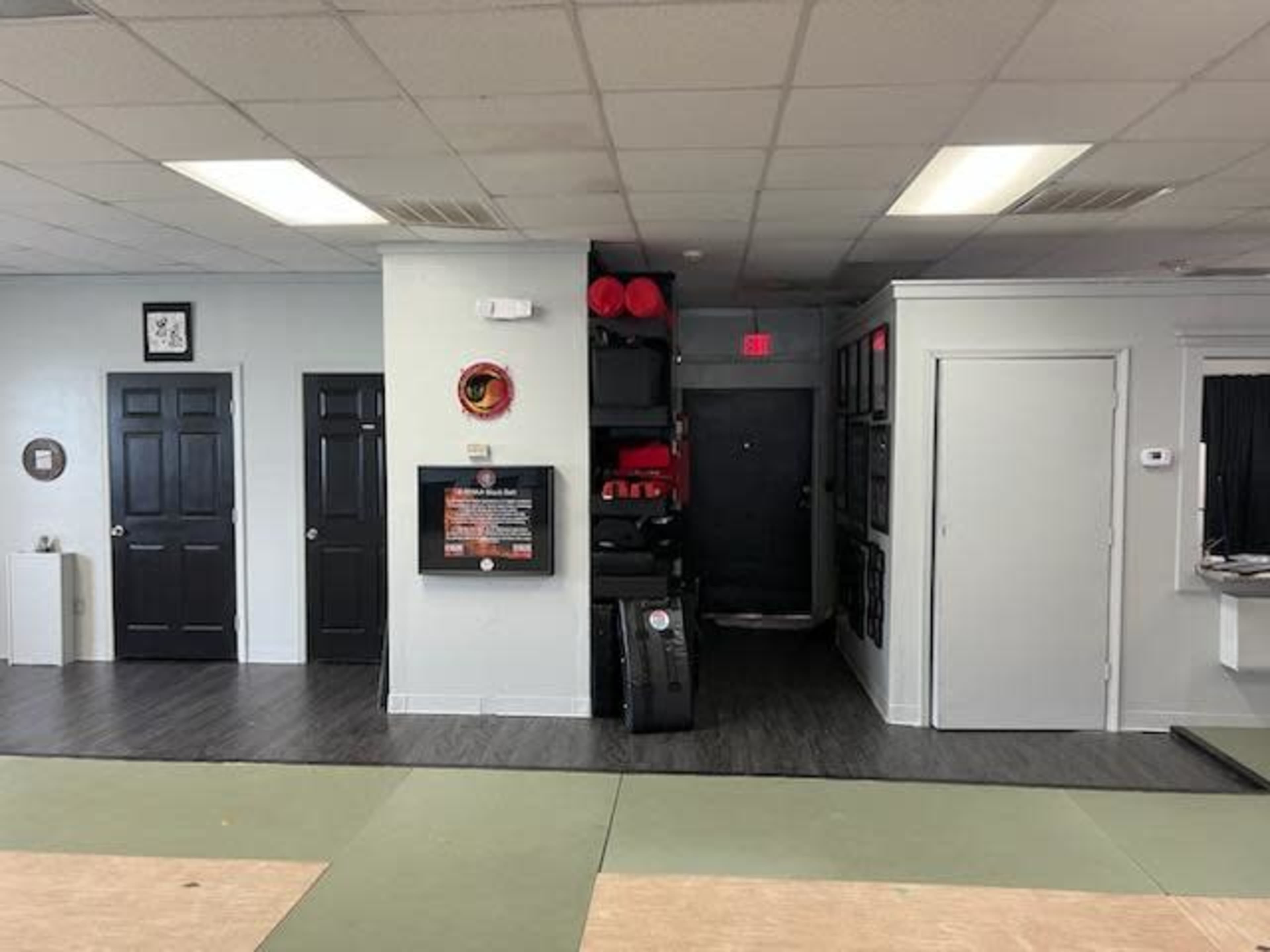 The image shows a training space with multiple doors on one side and stack of martial arts equipment, including red pads, against the wall on the other side.