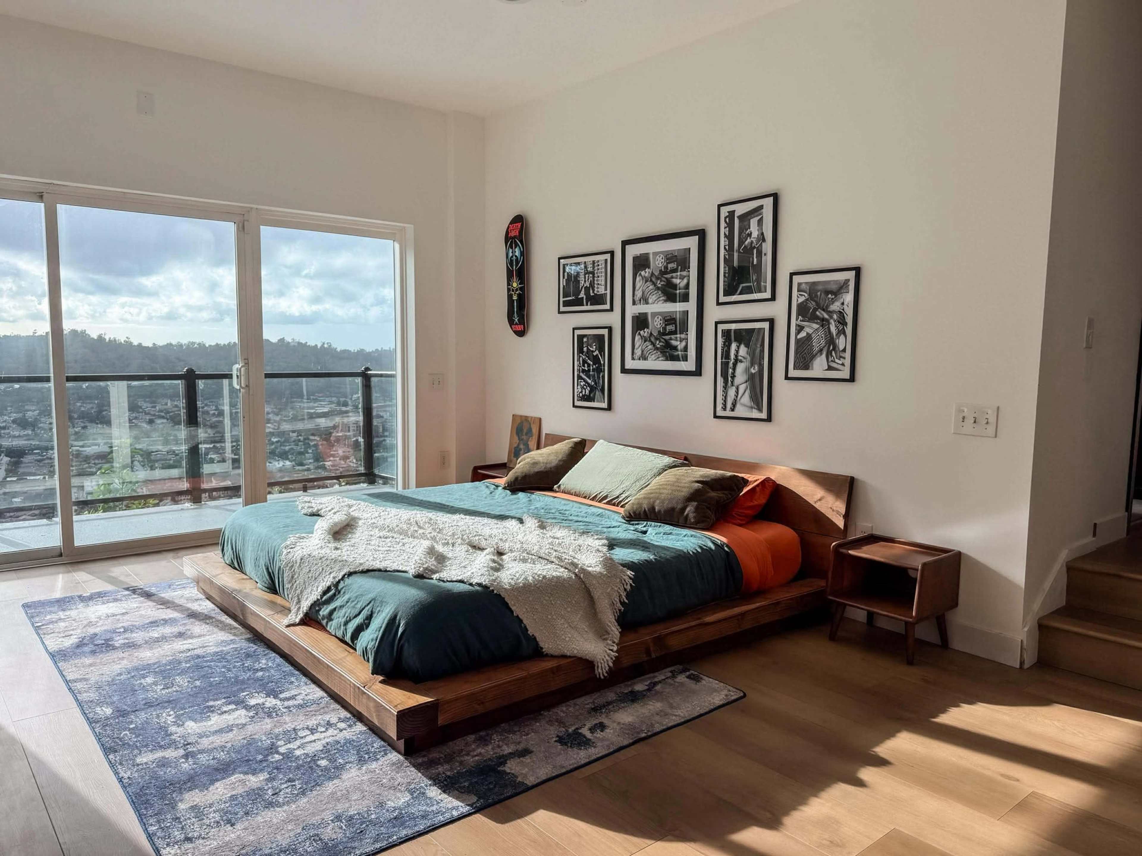 The image shows a modern bedroom with a large wooden bed, framed photographs on the wall, and a door leading to a balcony.