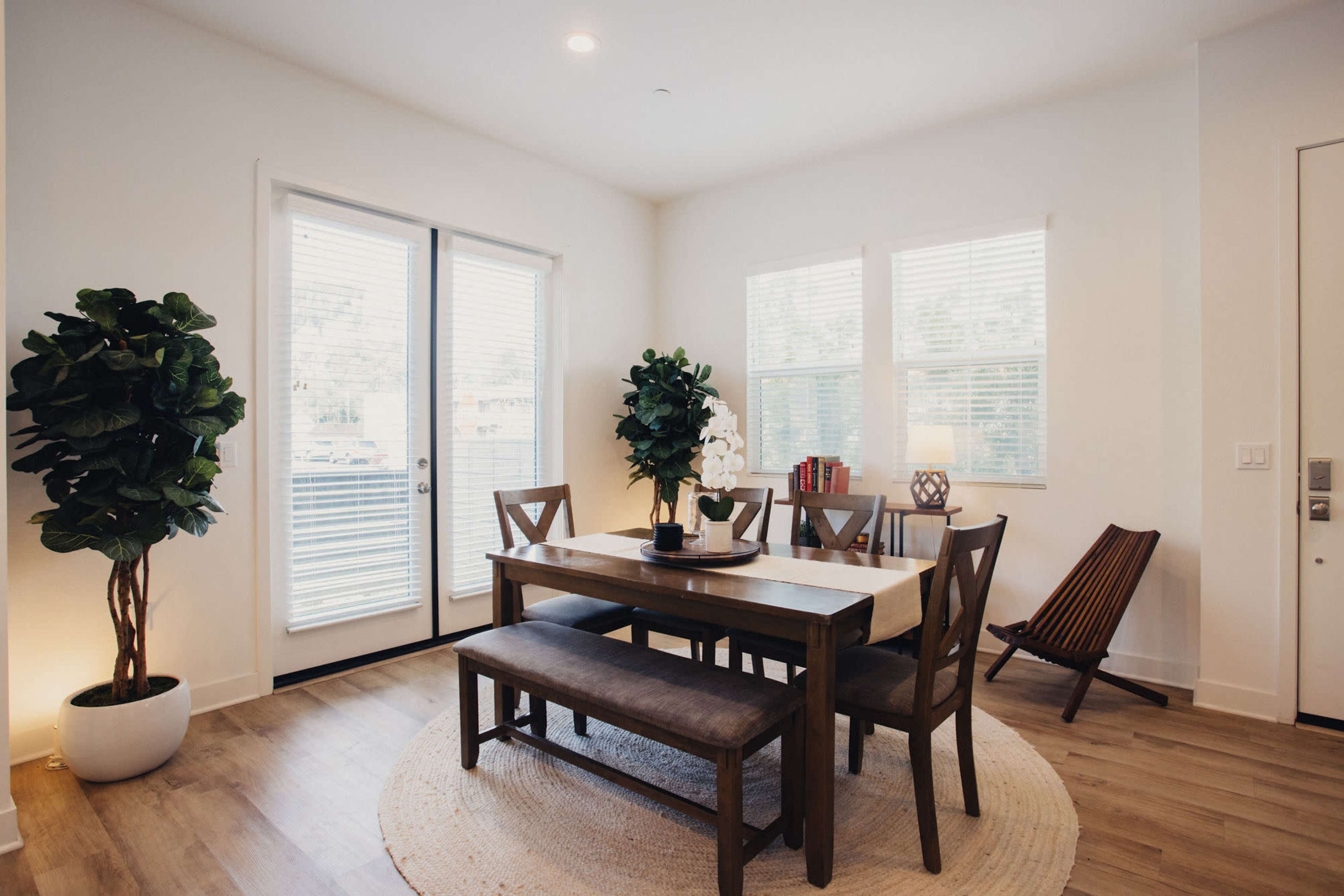 A dining area features a wooden table with benches, surrounded by chairs, a potted plant, and a small table with books and a lamp, all set near large windows.