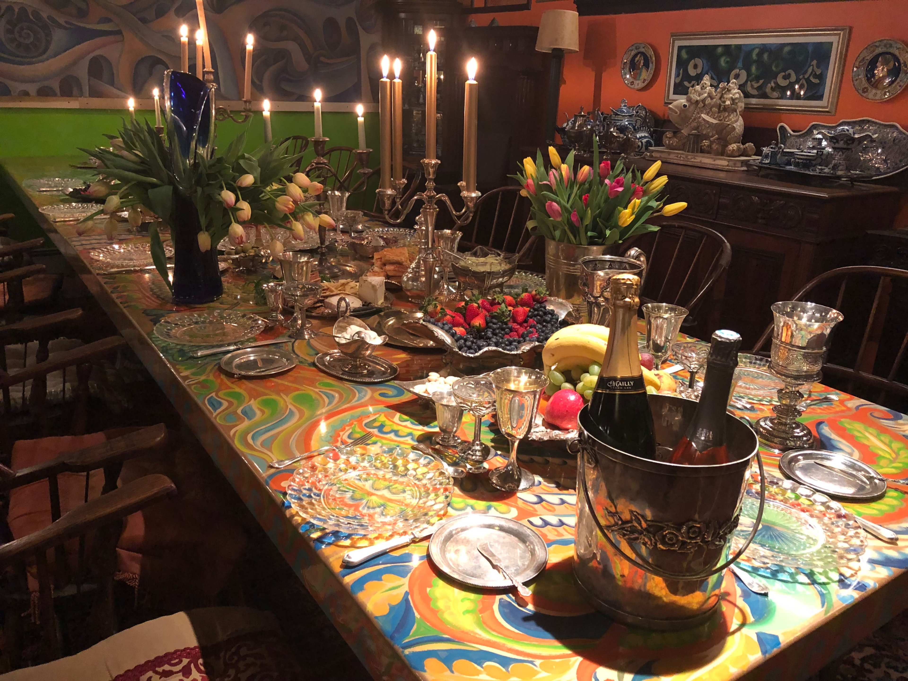 A long dining table is set with an array of dishes, fruits, and decorative centerpieces, illuminated by candles in silver candlesticks.