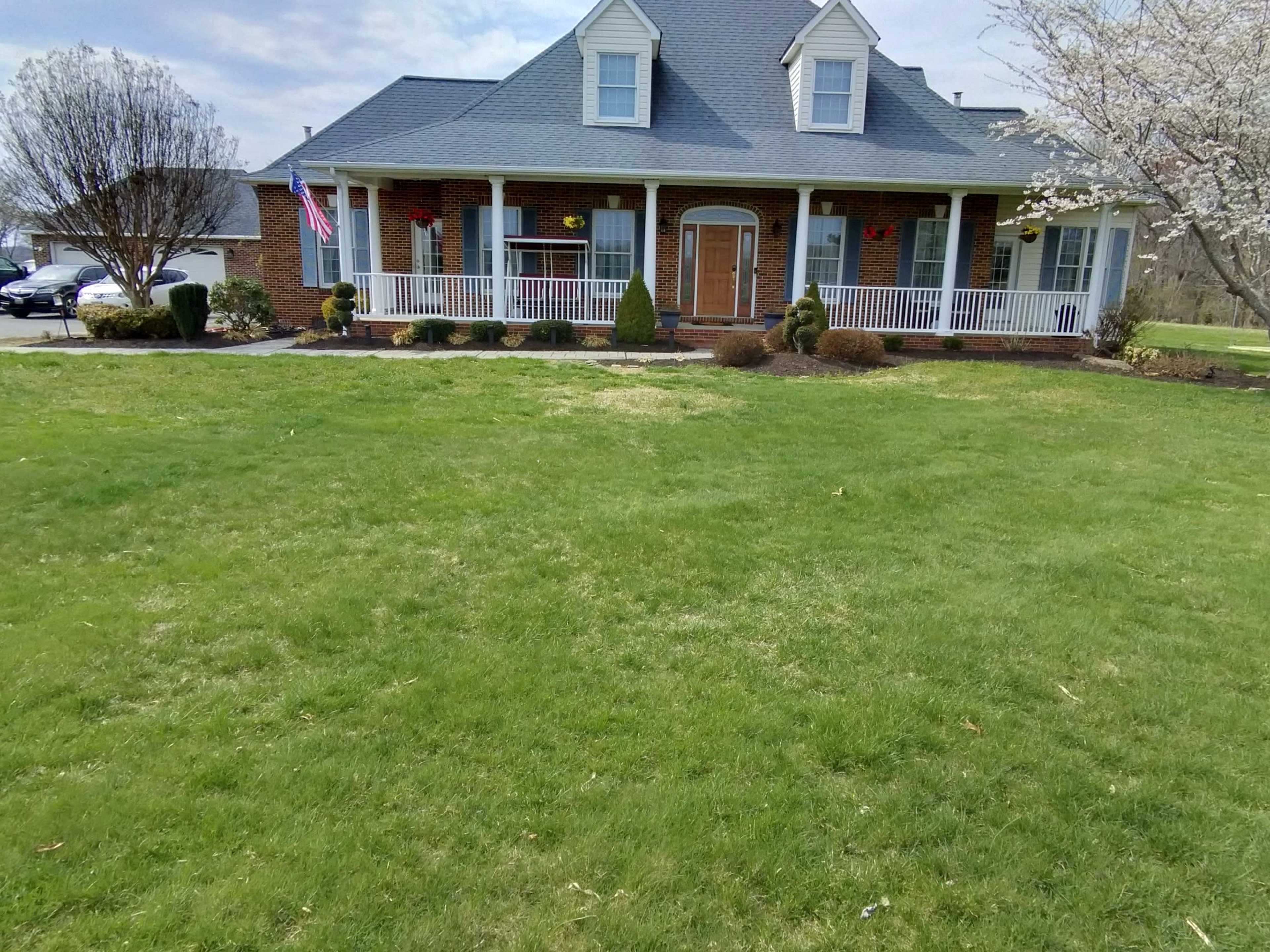 The image shows a single-story house with a front porch, a neatly trimmed lawn, and a driveway leading to parked vehicles.