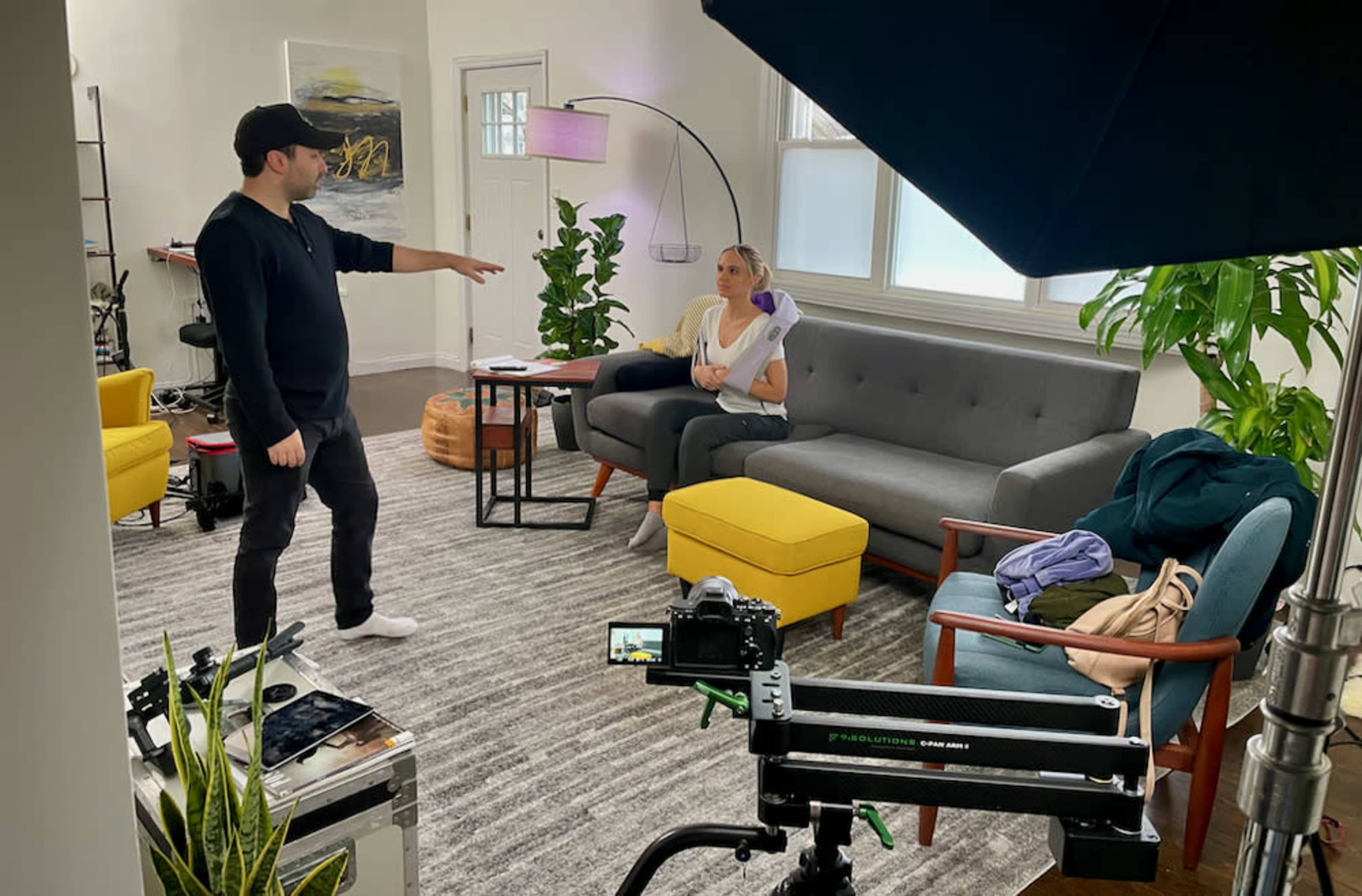 A man gestures while speaking to a woman seated on a gray couch in a well-lit living room with a mix of plants and furniture.