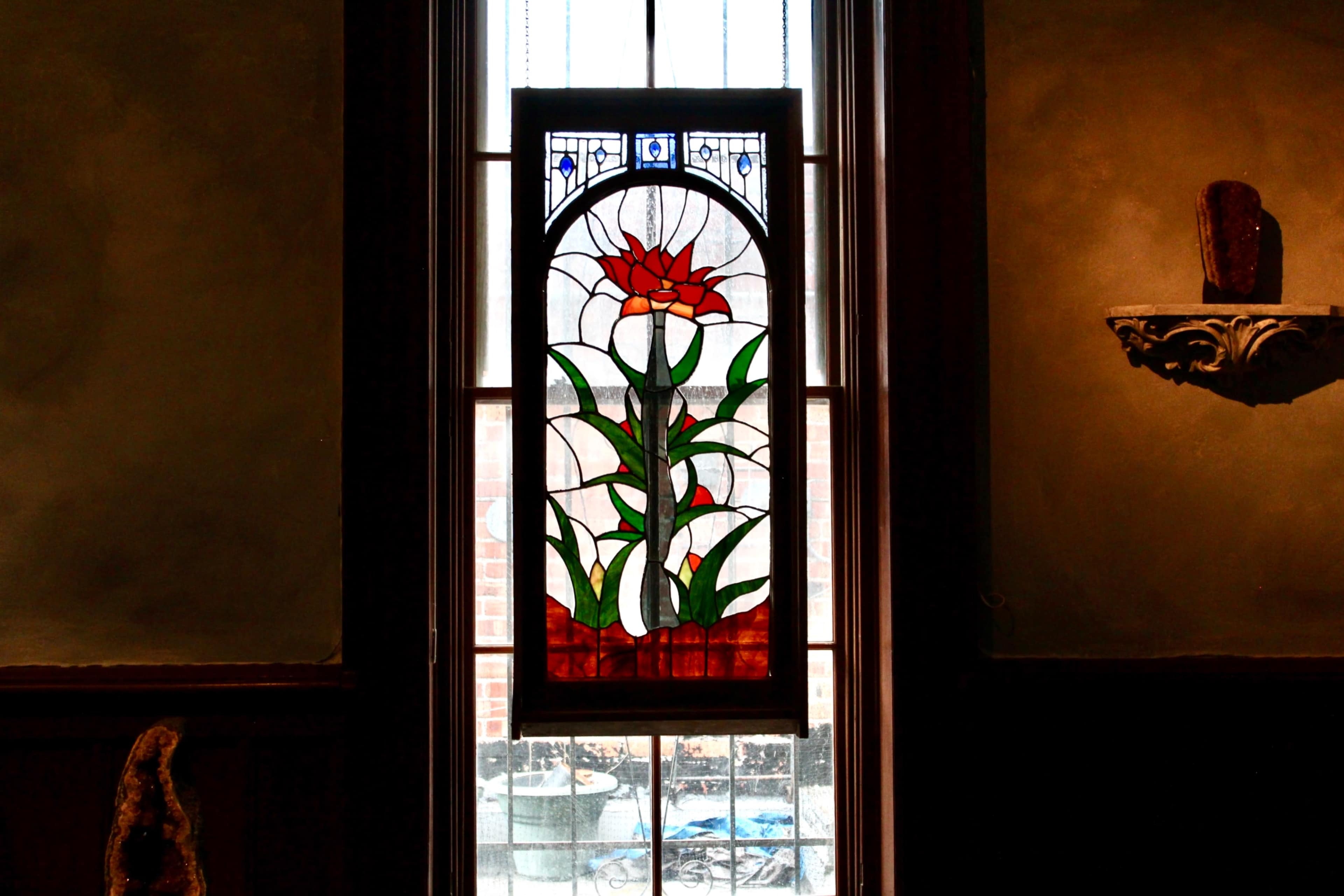 A stained glass window featuring a red flower and green leaves is positioned in front of a light source.