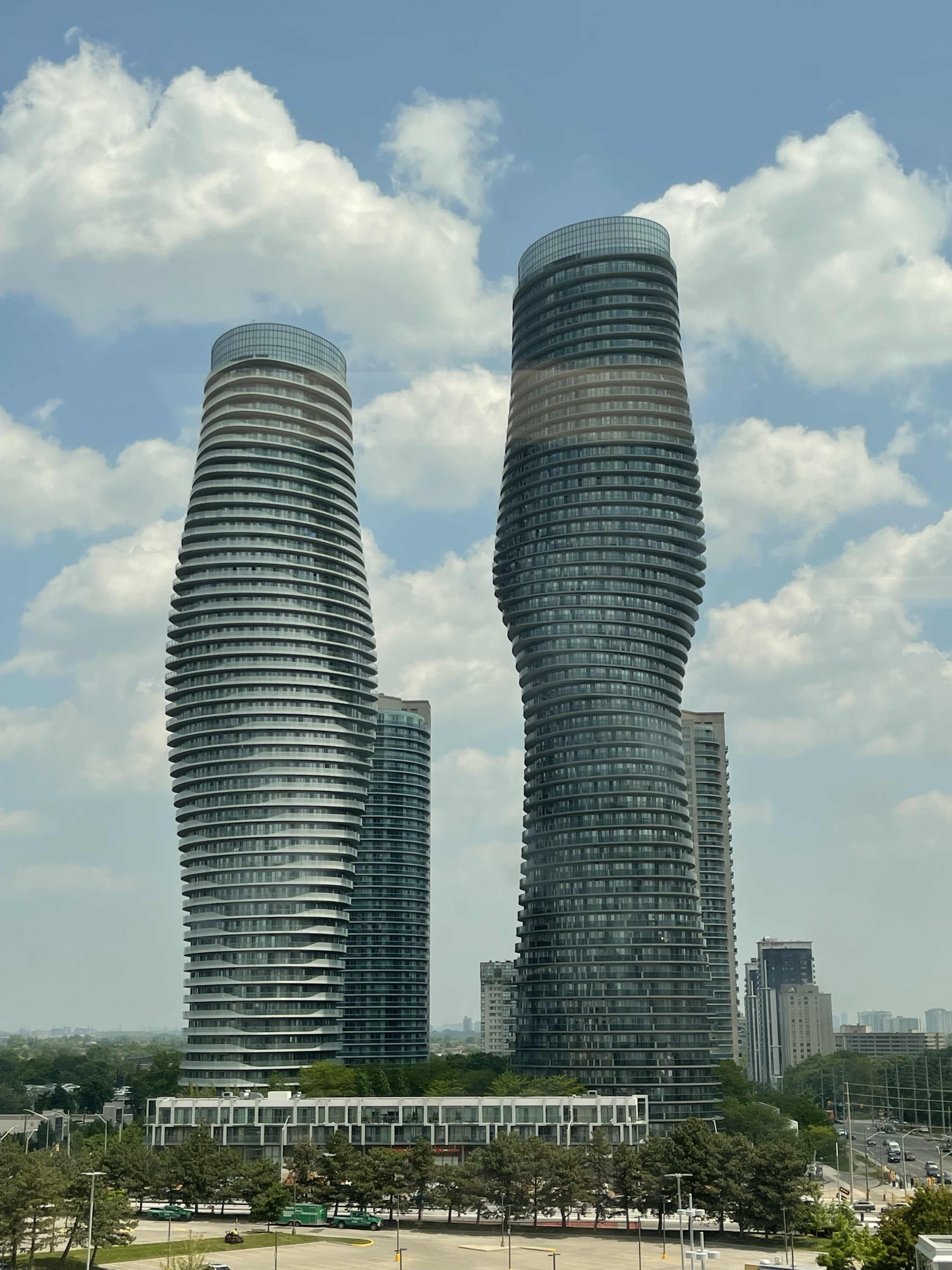 The image shows two modern, twisting skyscrapers rising against a partly cloudy blue sky in an urban area.