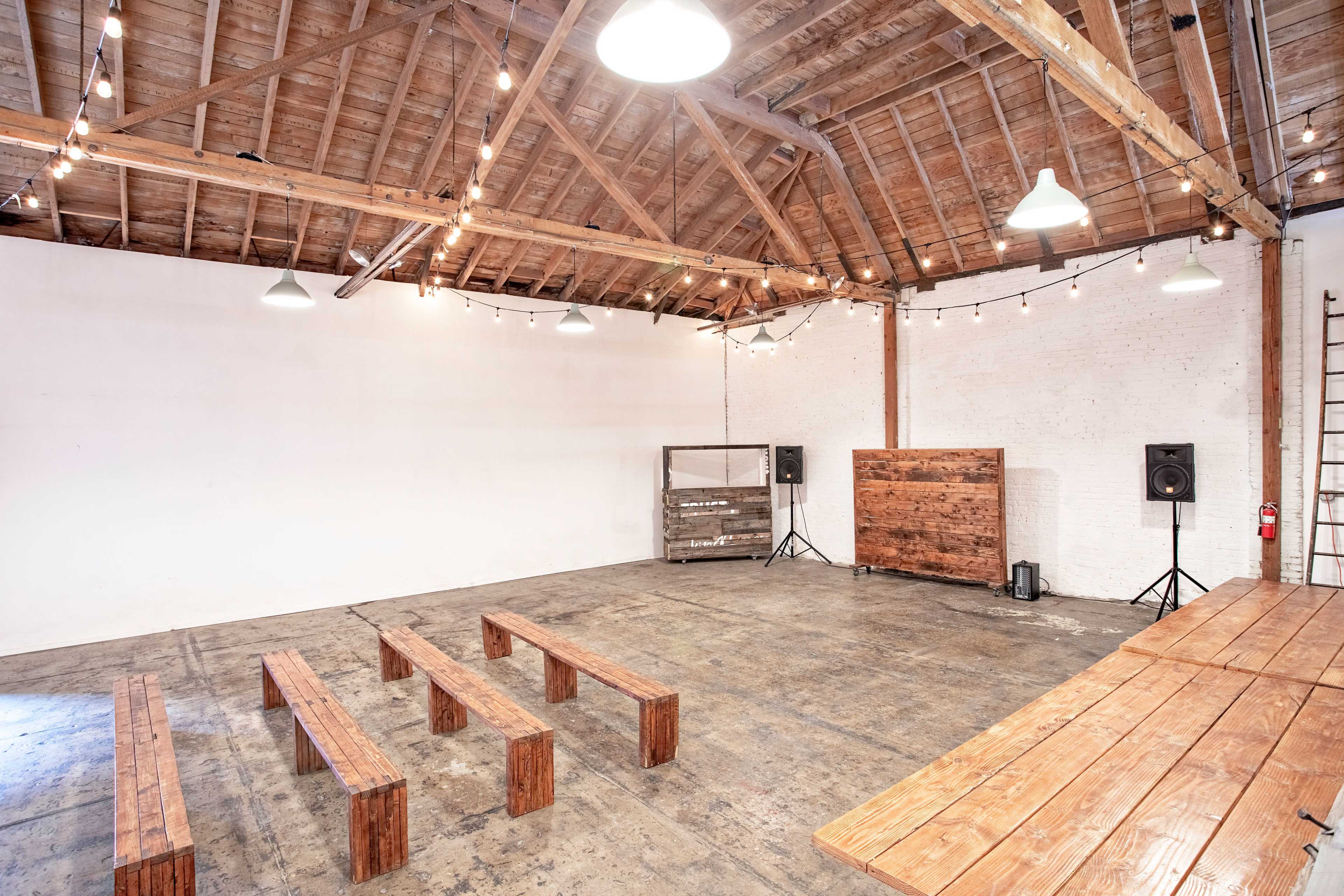 A spacious event venue with wooden benches, a long table, and minimalist decor under a ceiling with exposed beams and string lights.