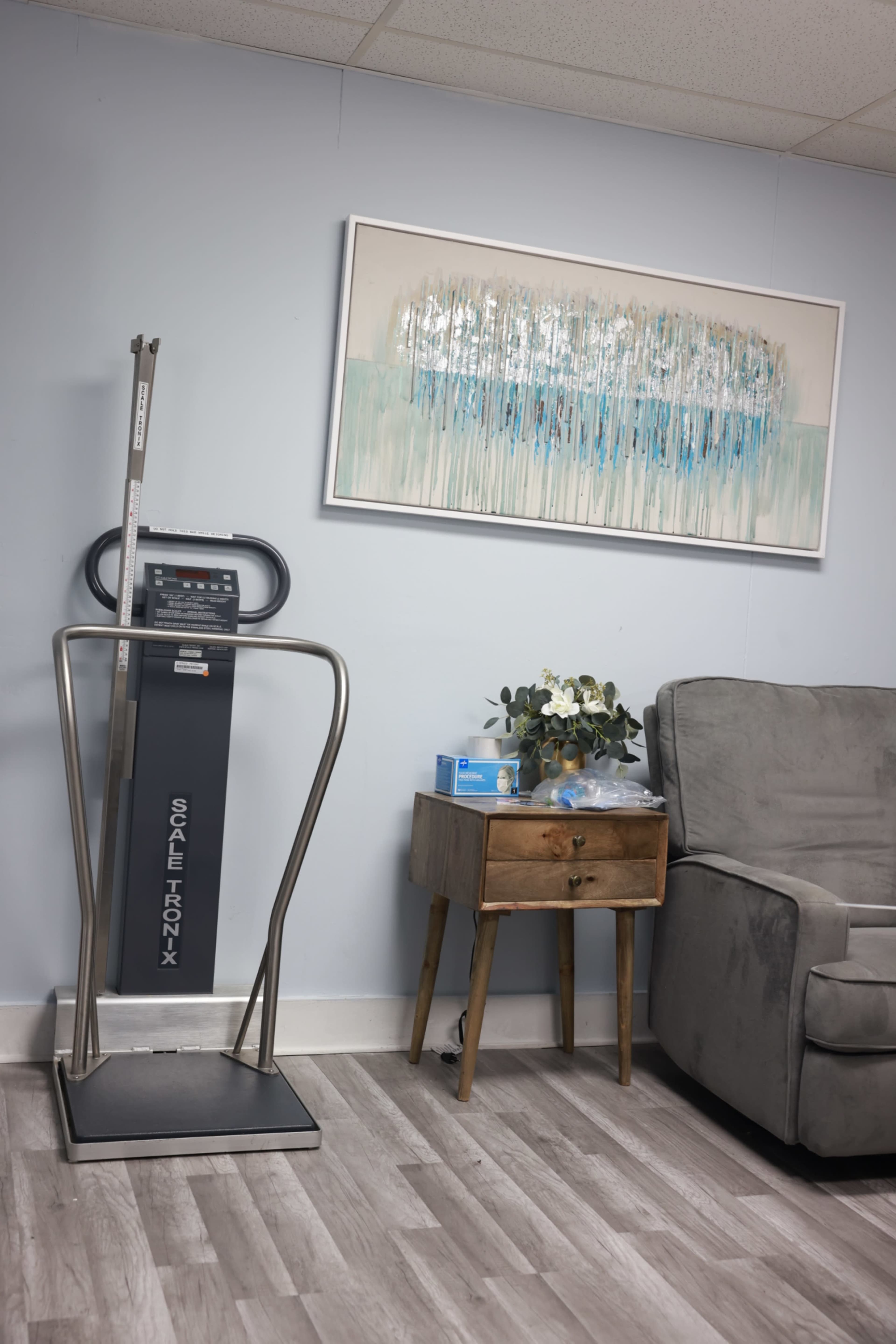The image shows a medical weighing scale next to a small wooden side table with flowers and medical supplies, positioned in a room with a couch and abstract artwork on the wall.