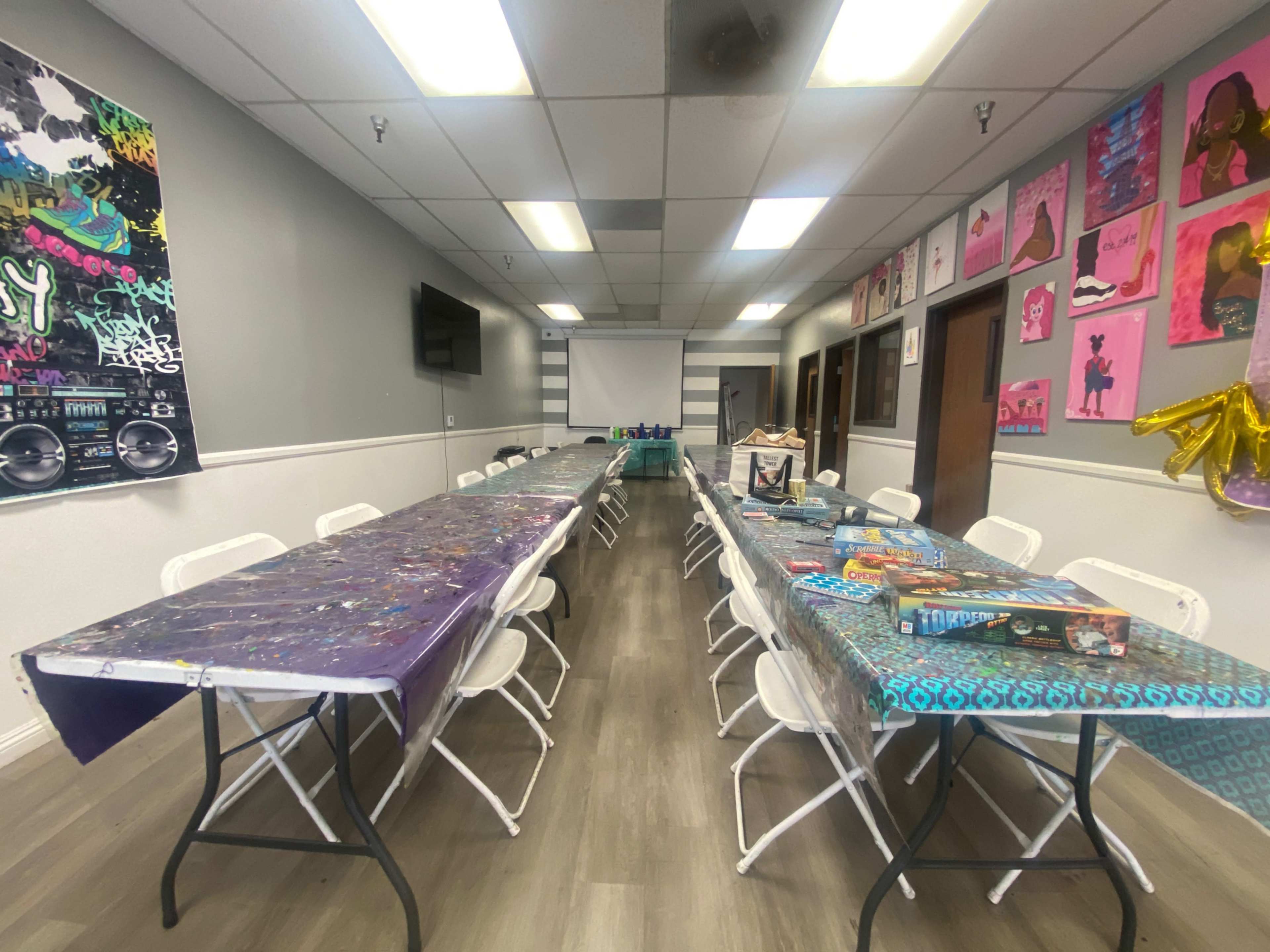 The image shows an elongated room with several tables covered in colorful tablecloths and chairs, decorated with artwork on the walls and a screen at the end.