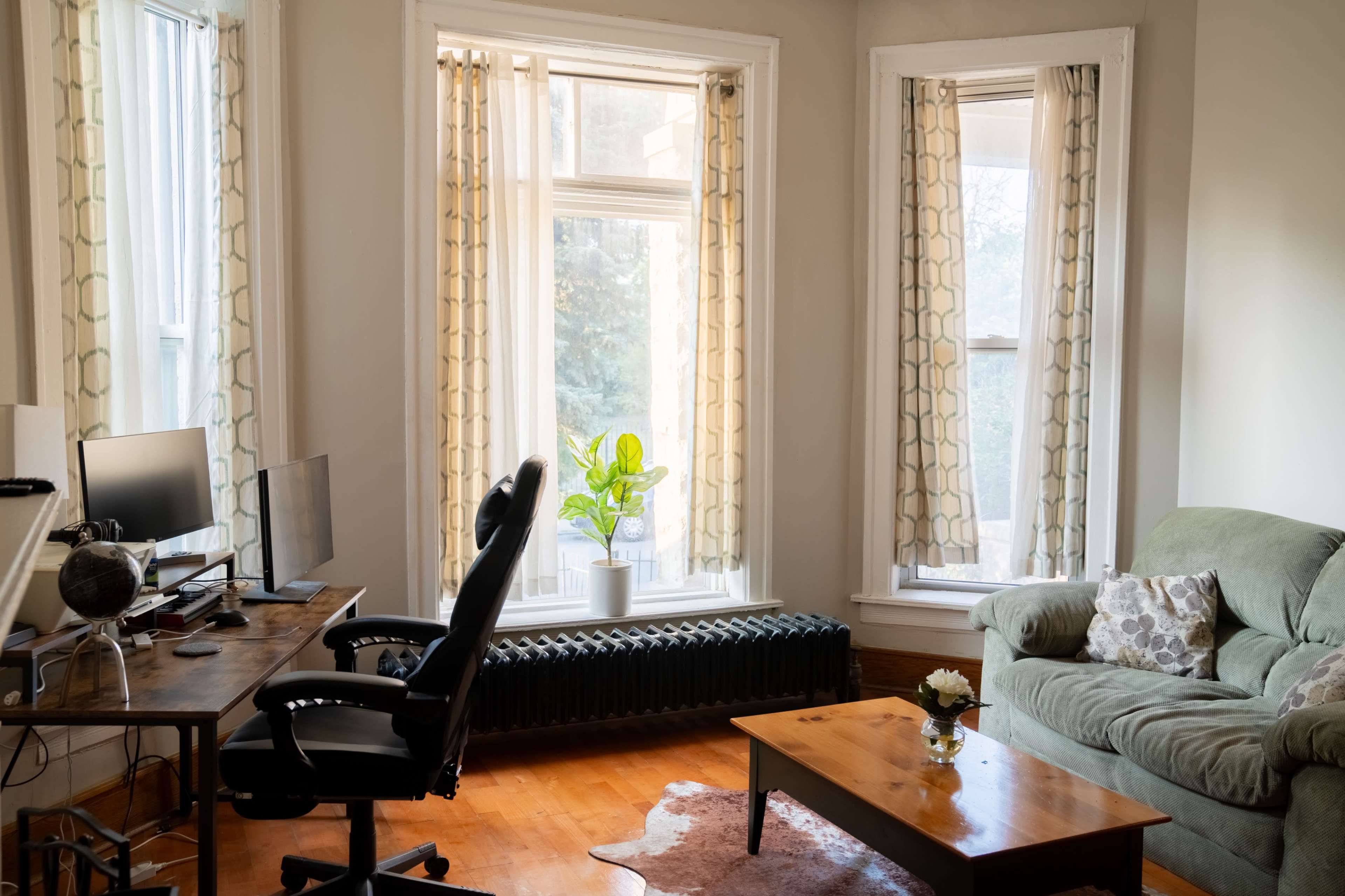 A living room features a green sofa, a wooden coffee table, a desk with a computer setup, and large windows with light curtains, along with a potted plant in front of the window.