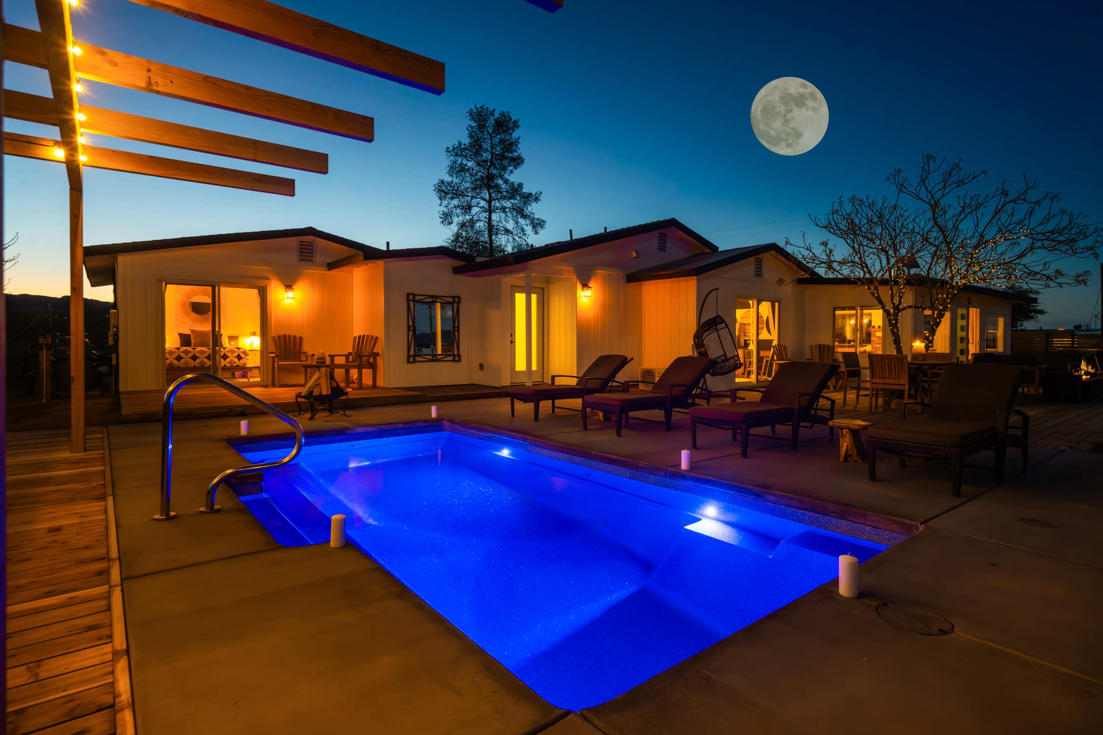 A modern home with a swimming pool is illuminated at dusk, featuring lounge chairs and a full moon in the sky.