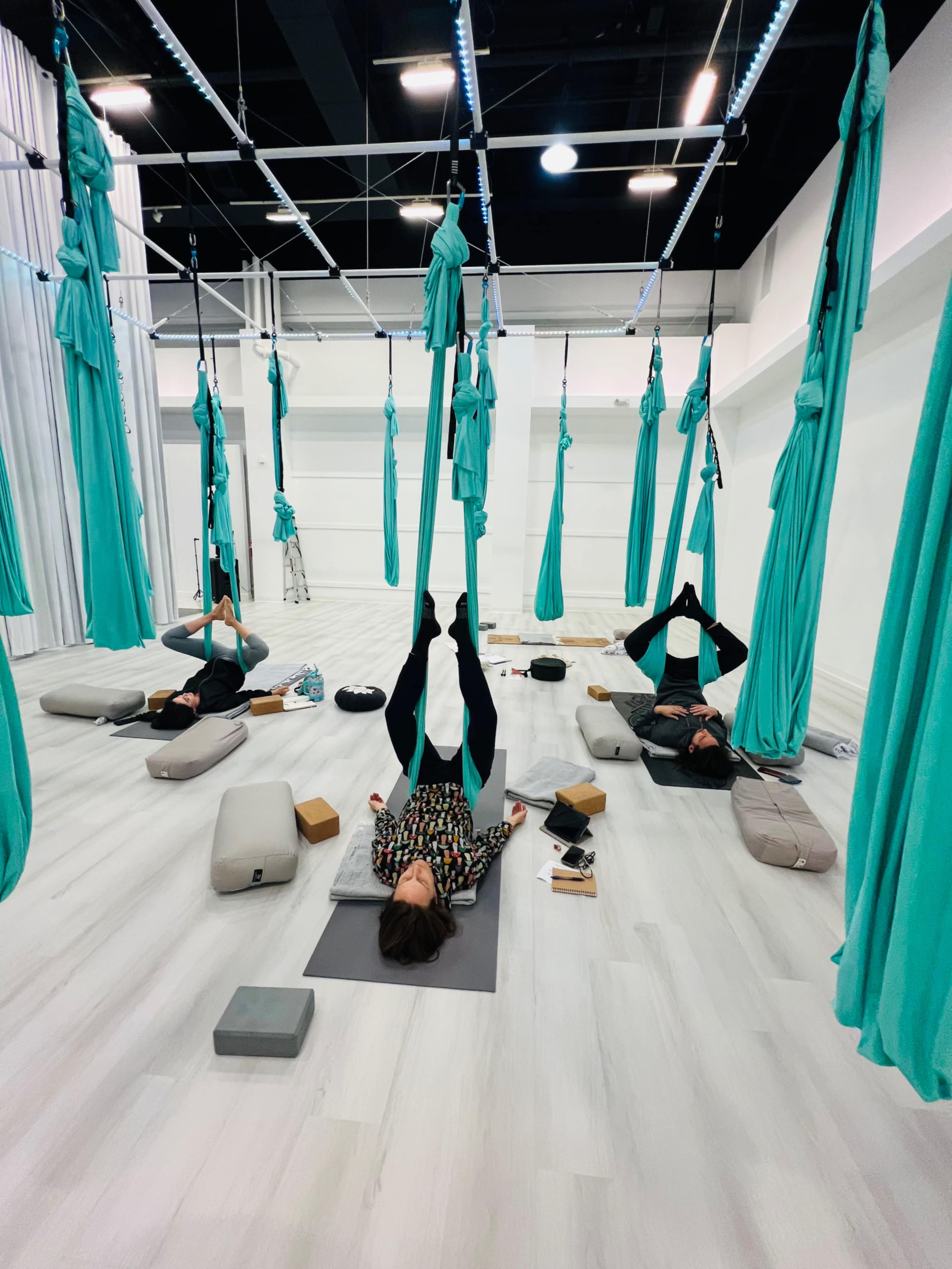 A group of people is practicing aerial yoga, hanging upside down in teal hammocks in a spacious, well-lit studio.