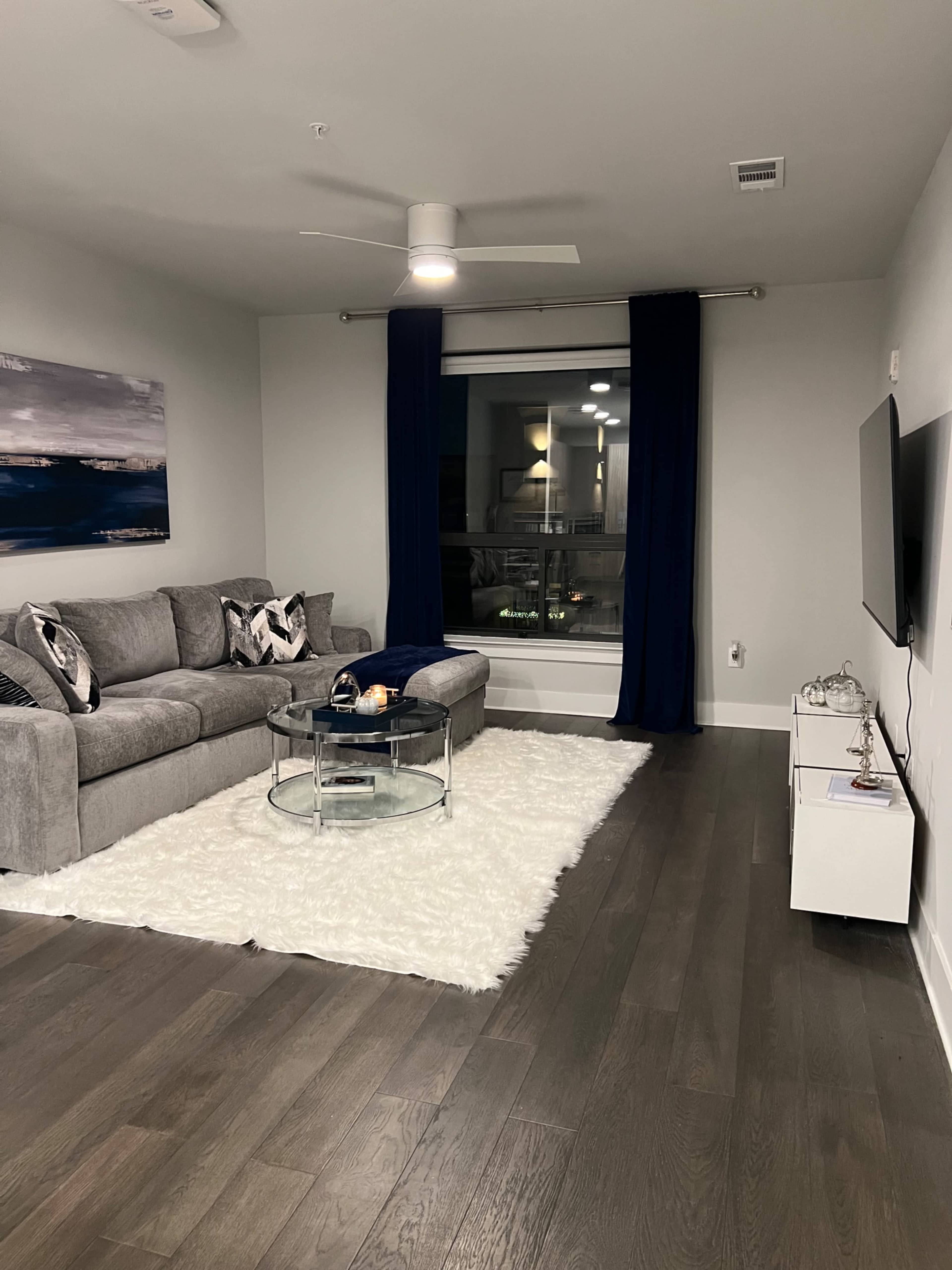 A modern living room with a gray sectional sofa, a glass coffee table on a white rug, and large windows dressed with dark blue curtains.