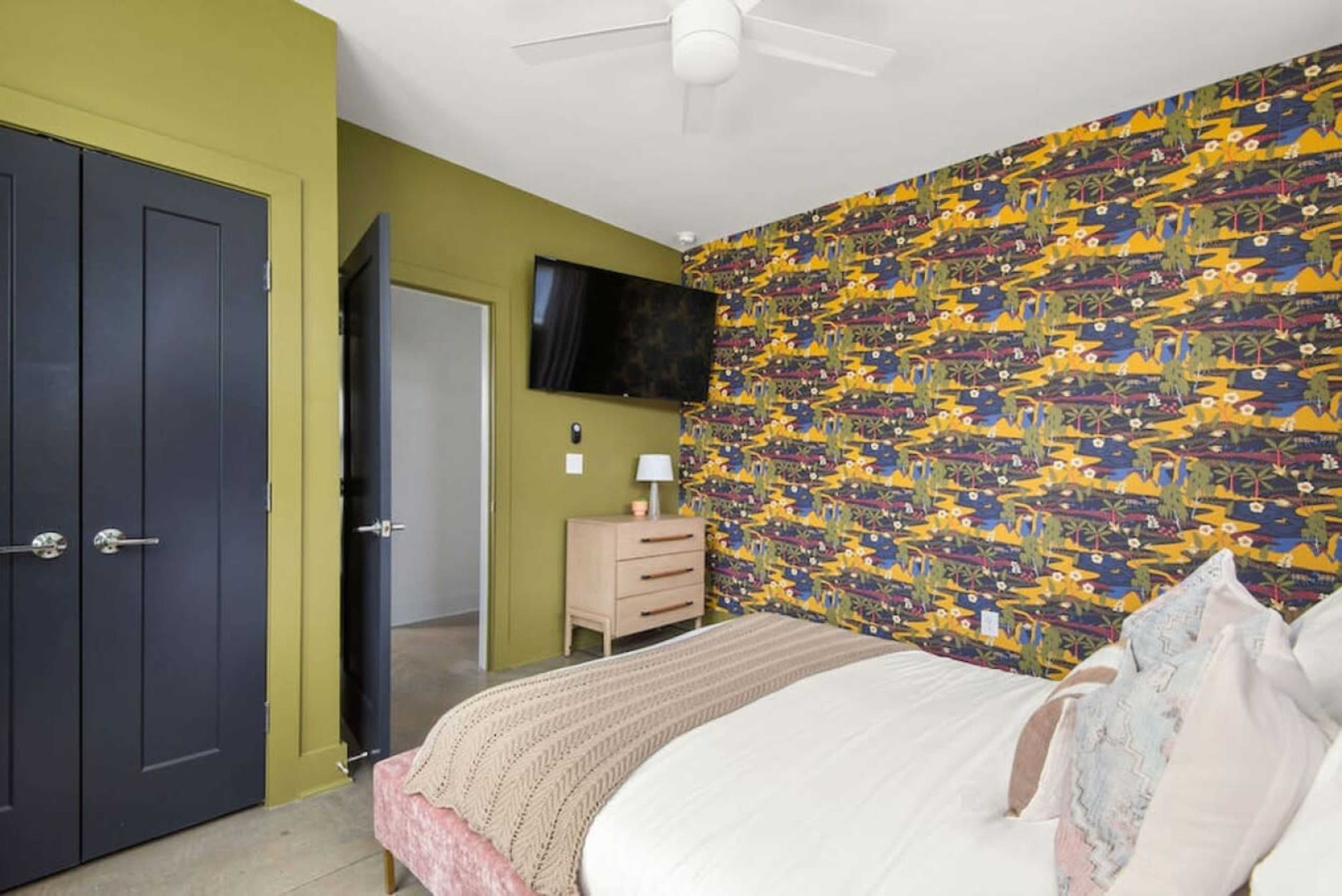 A bedroom with a green wall, patterned wallpaper, a bed with a knit throw, a low wooden nightstand, and a mounted television.