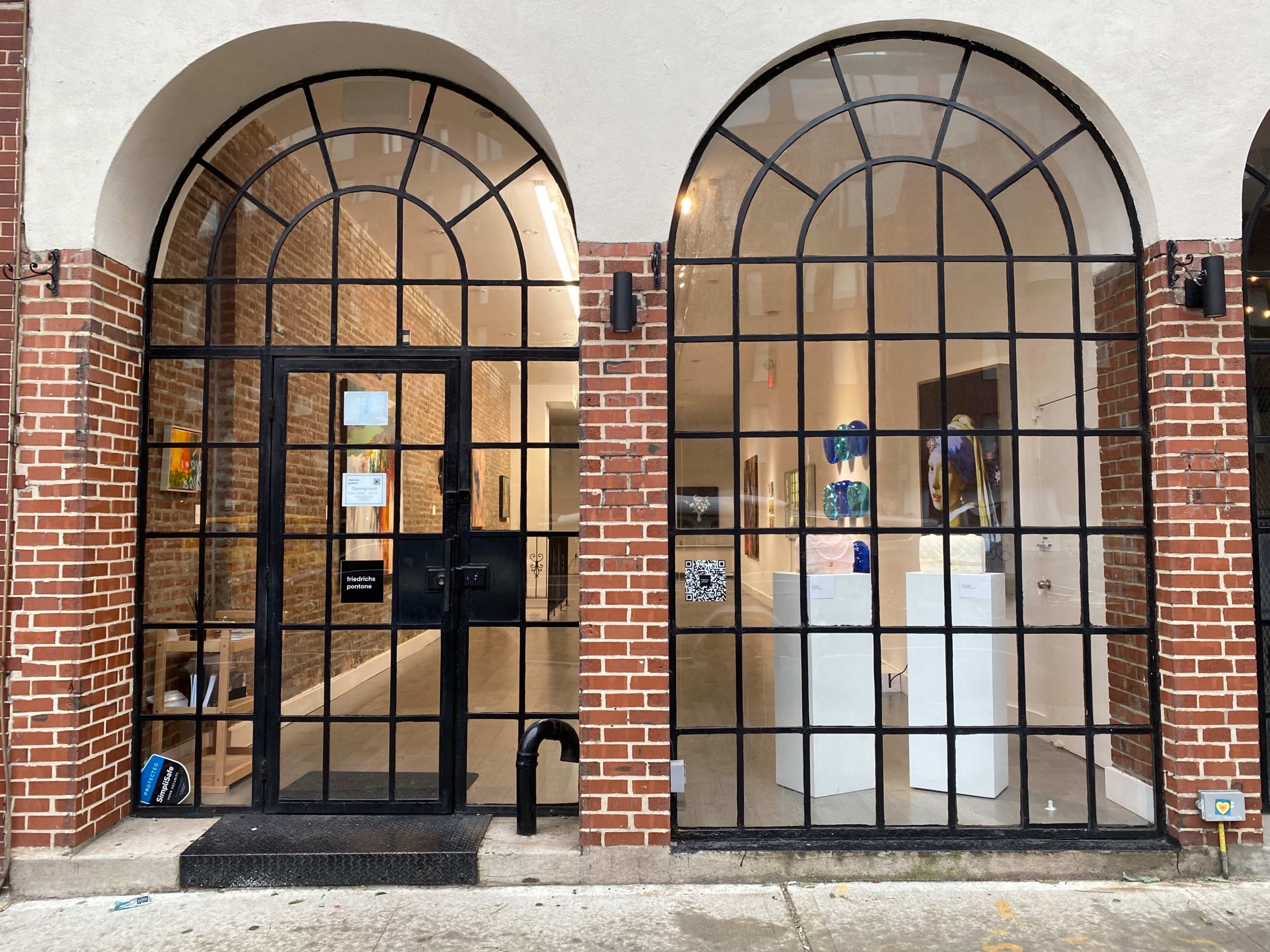 The image shows a building facade with two large arched windows flanking a black entrance door, revealing an art gallery inside.