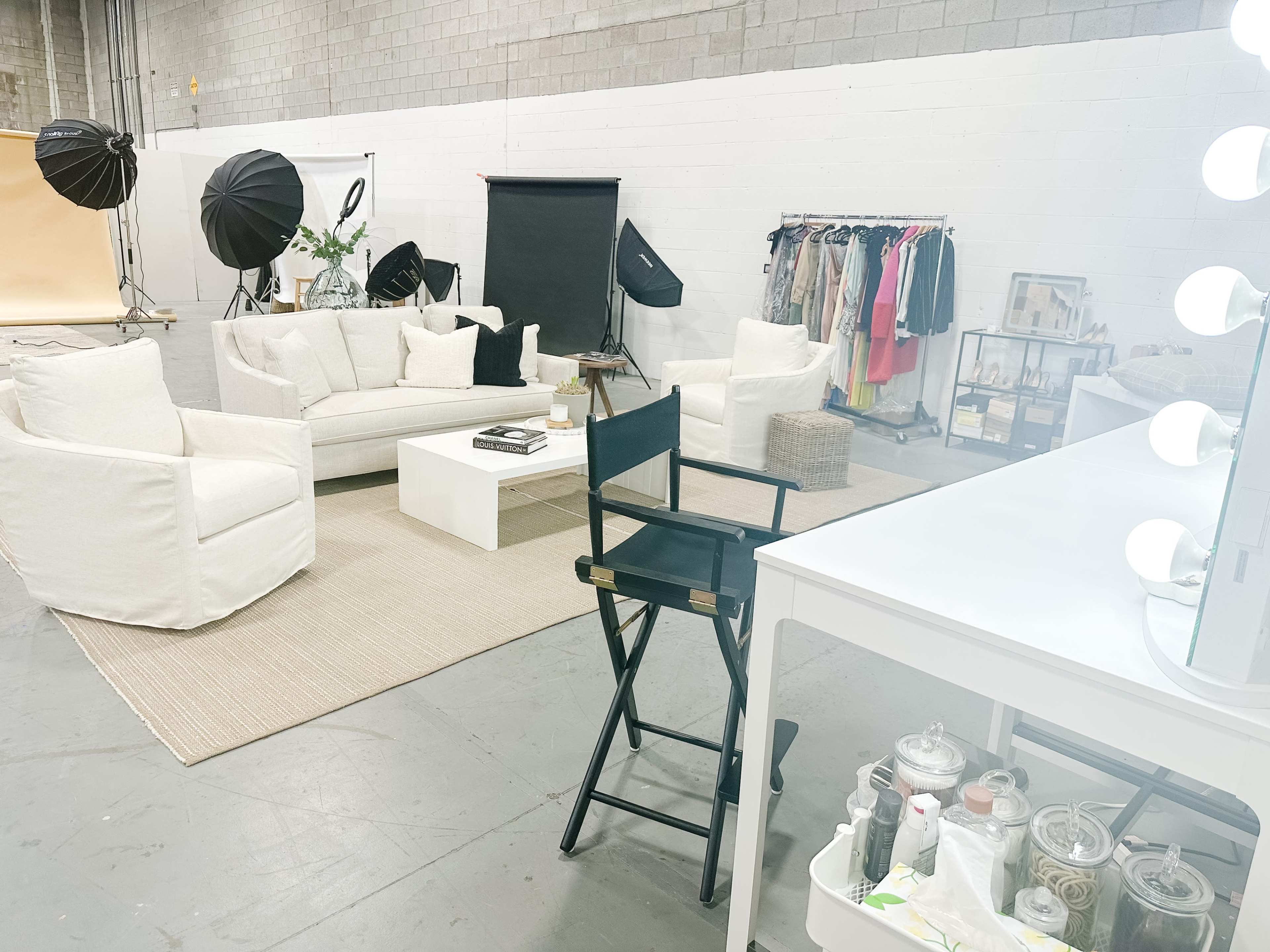 A spacious studio features white seating, a coffee table, a clothing rack, and a vanity mirror with lights, surrounded by photography equipment.