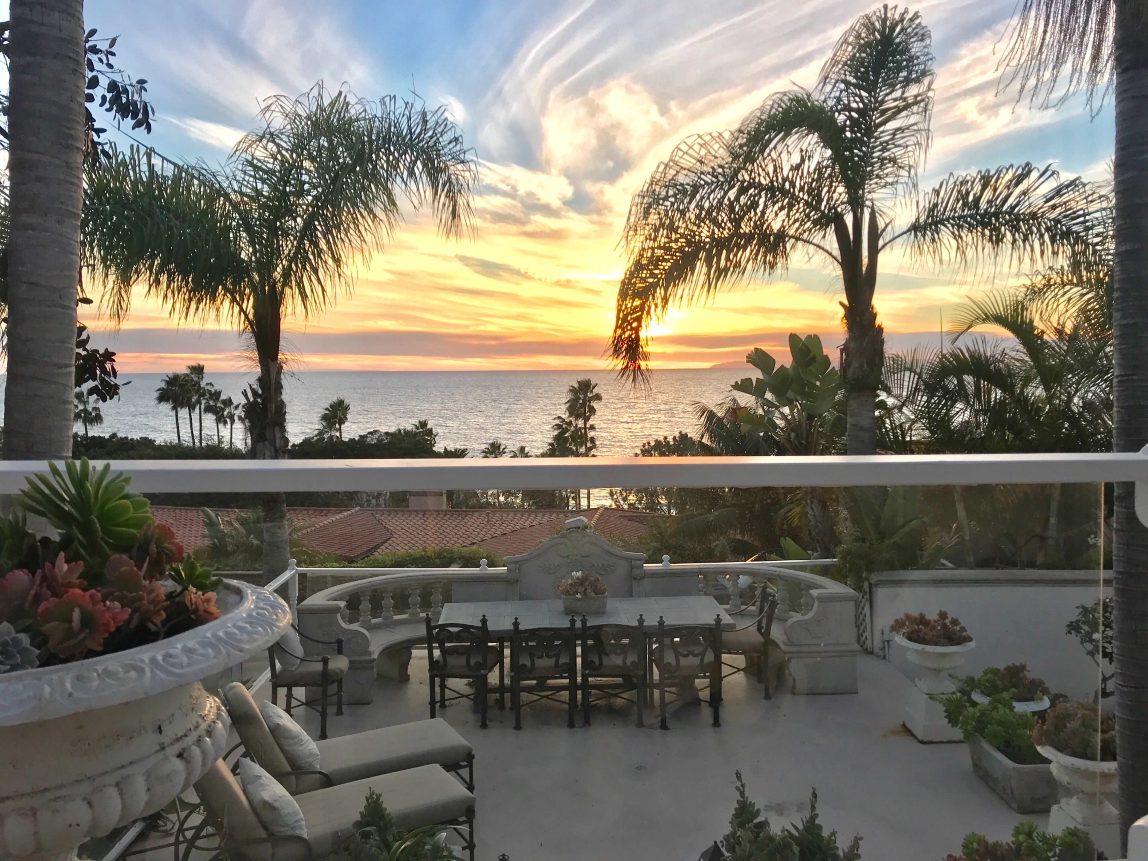 The image features a patio with a large dining table and chairs, overlooking a sunset over the ocean framed by palm trees.