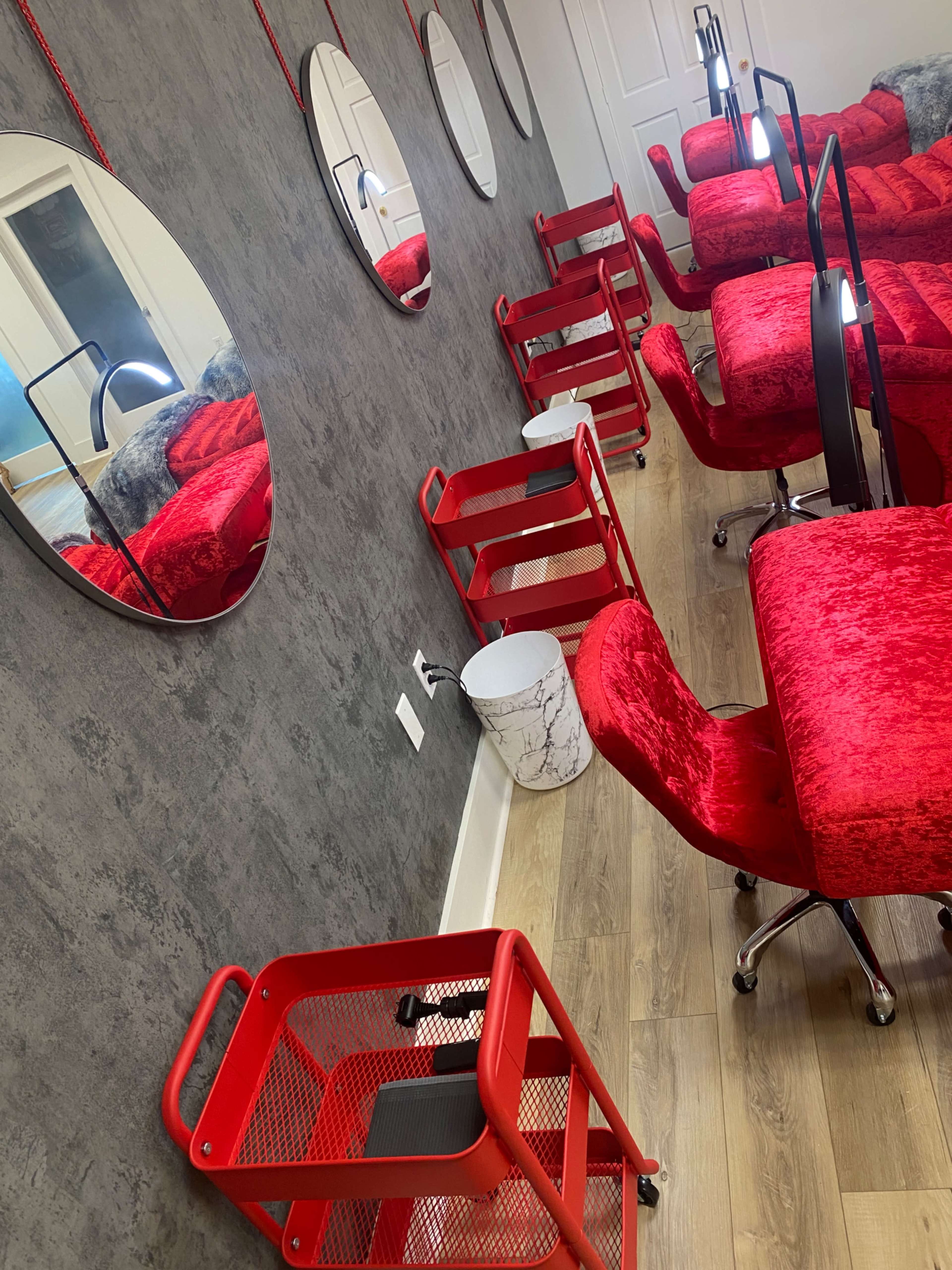 The image shows a salon interior with several red chairs, circular mirrors on the wall, and red mobile carts arranged on a wooden floor.
