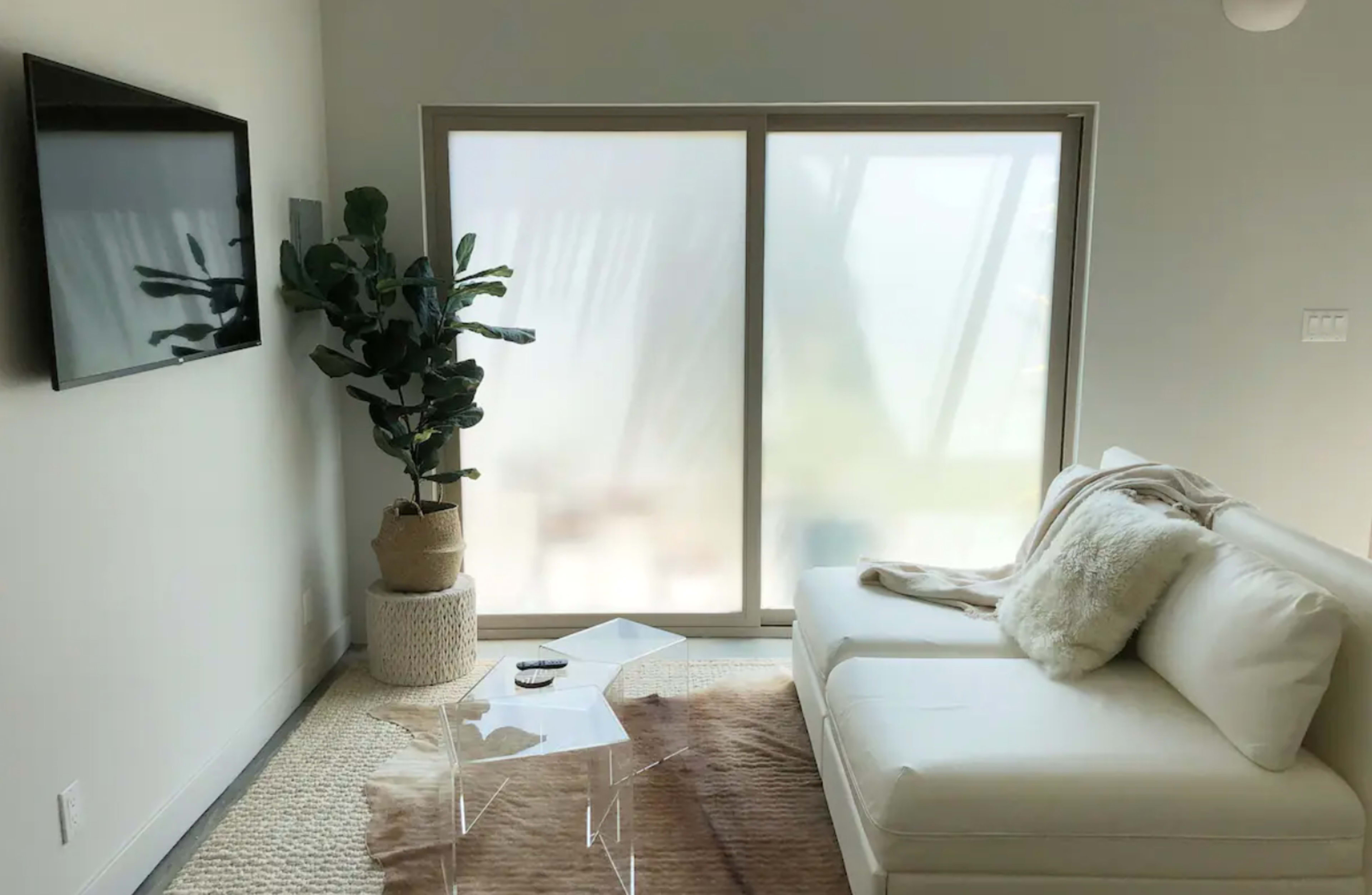 A bright living room features a white sectional sofa, a clear coffee table, and a large window with sheer curtains.
