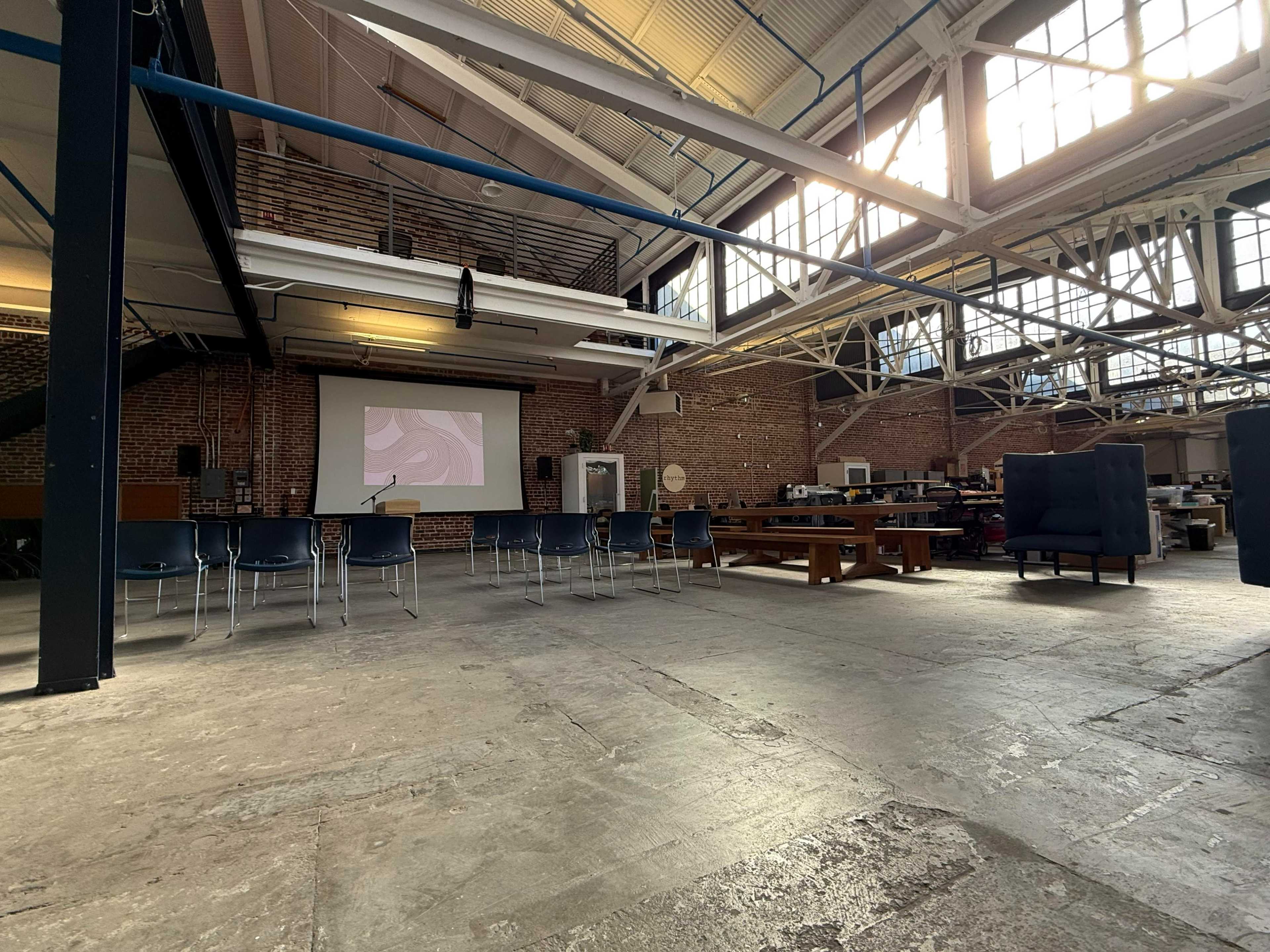 The image shows a spacious industrial interior with high ceilings, large windows, and a projection screen set up in front of several chairs.